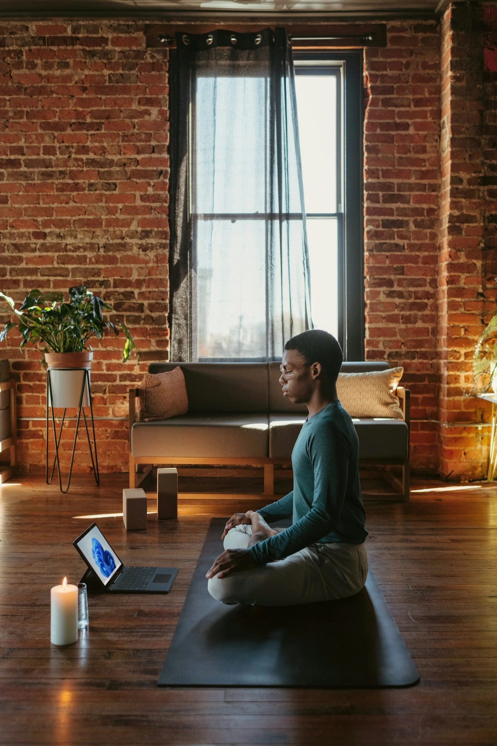 A Black man meditating in lotus pose on a mat in a brick-wall Brooklyn loft with a candle and laptop