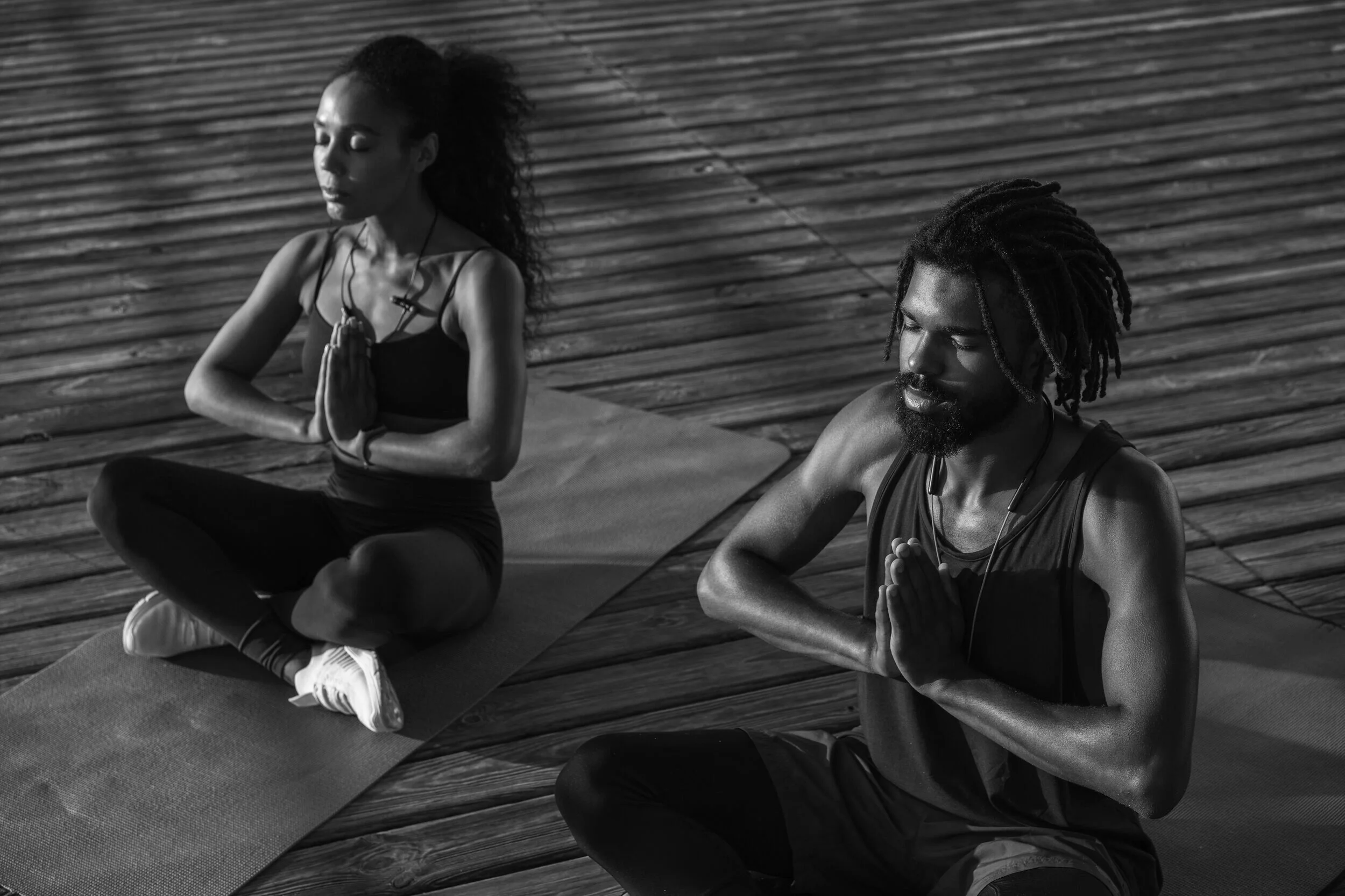 A Black man and woman seated cross-legged in meditation with hands in prayer on a wooden deck