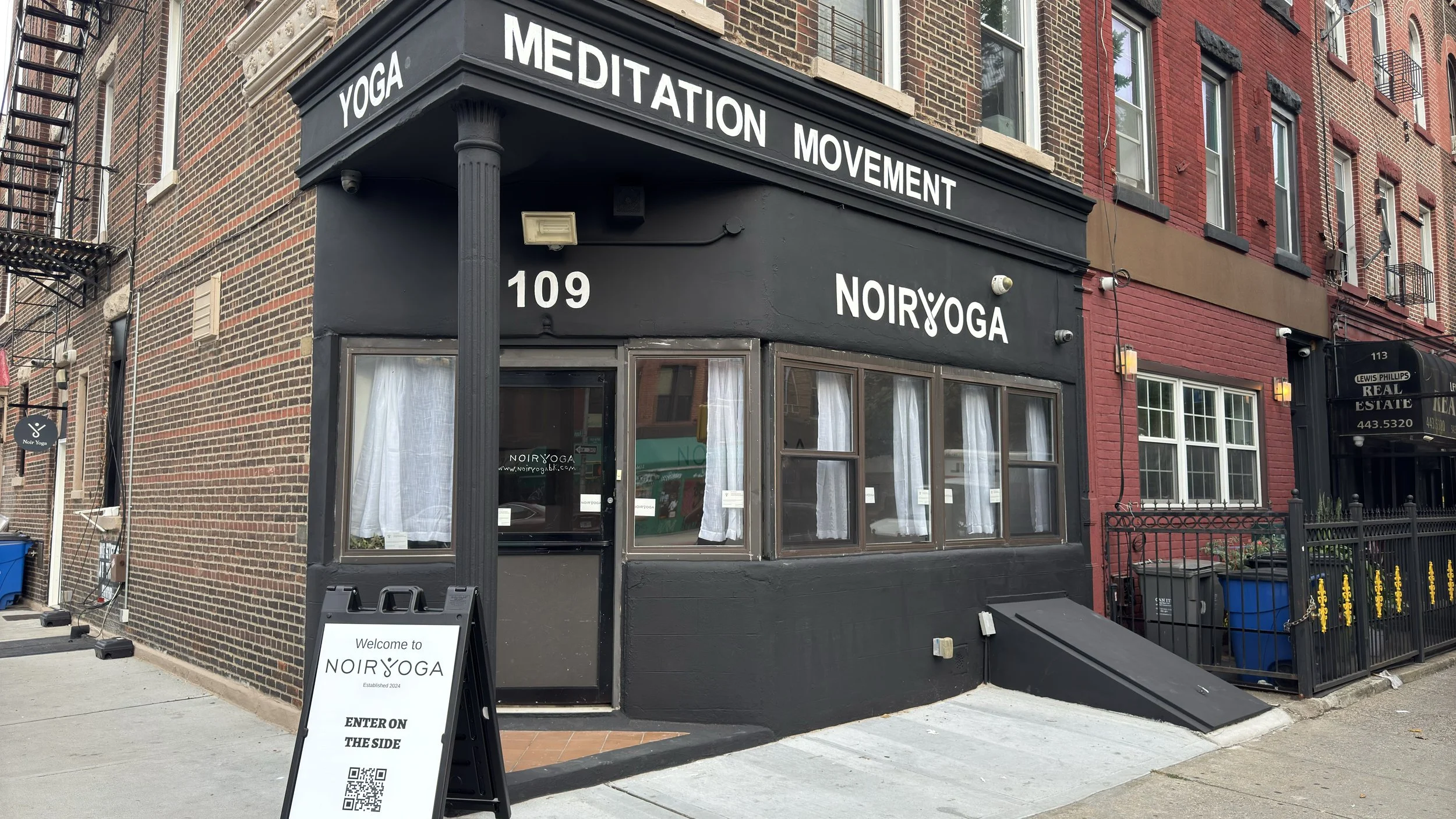 The outside of Noir Yoga at 109 Ralph Avenue, white letters reading Yoga, Meditation, Movement on a black storefront