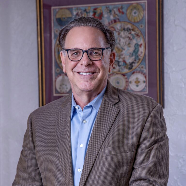 Portrait of Dr. Jeffrey S. Block in 2023, wearing a grey jacket and blue shirt, framed artwork behind him