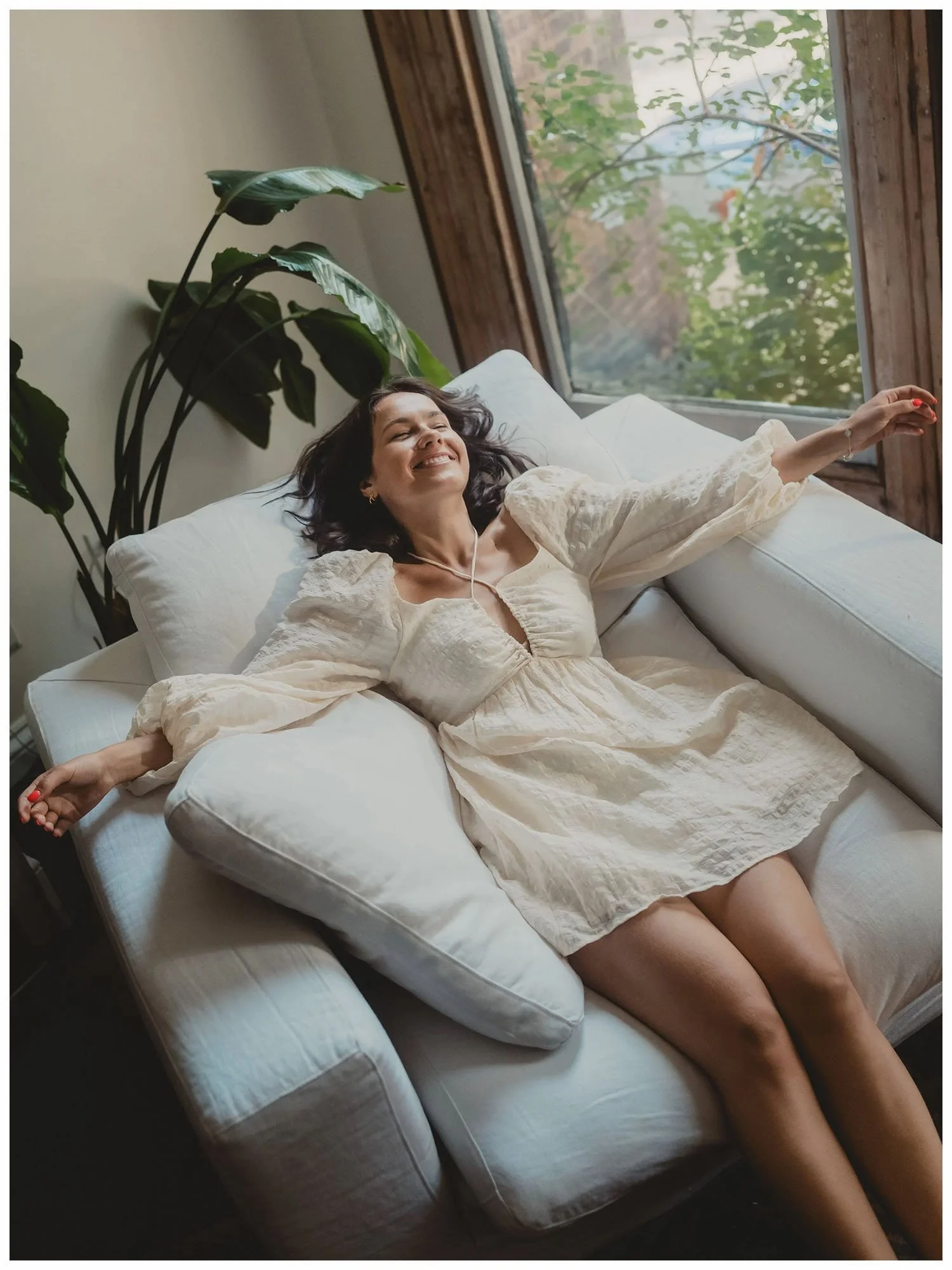 Woman stretching contentedly on a white couch beside a sunlit window