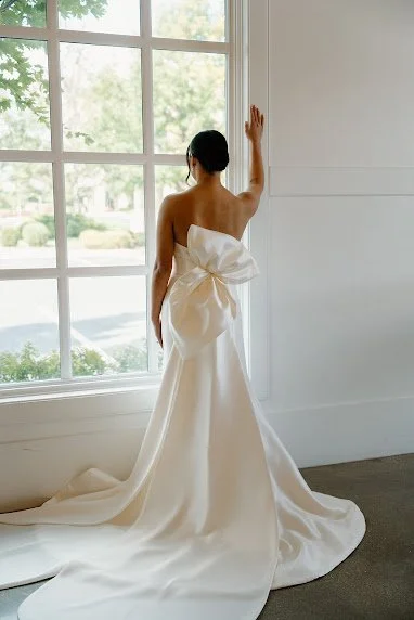 Bride in a satin bow gown at a window overlooking a garden