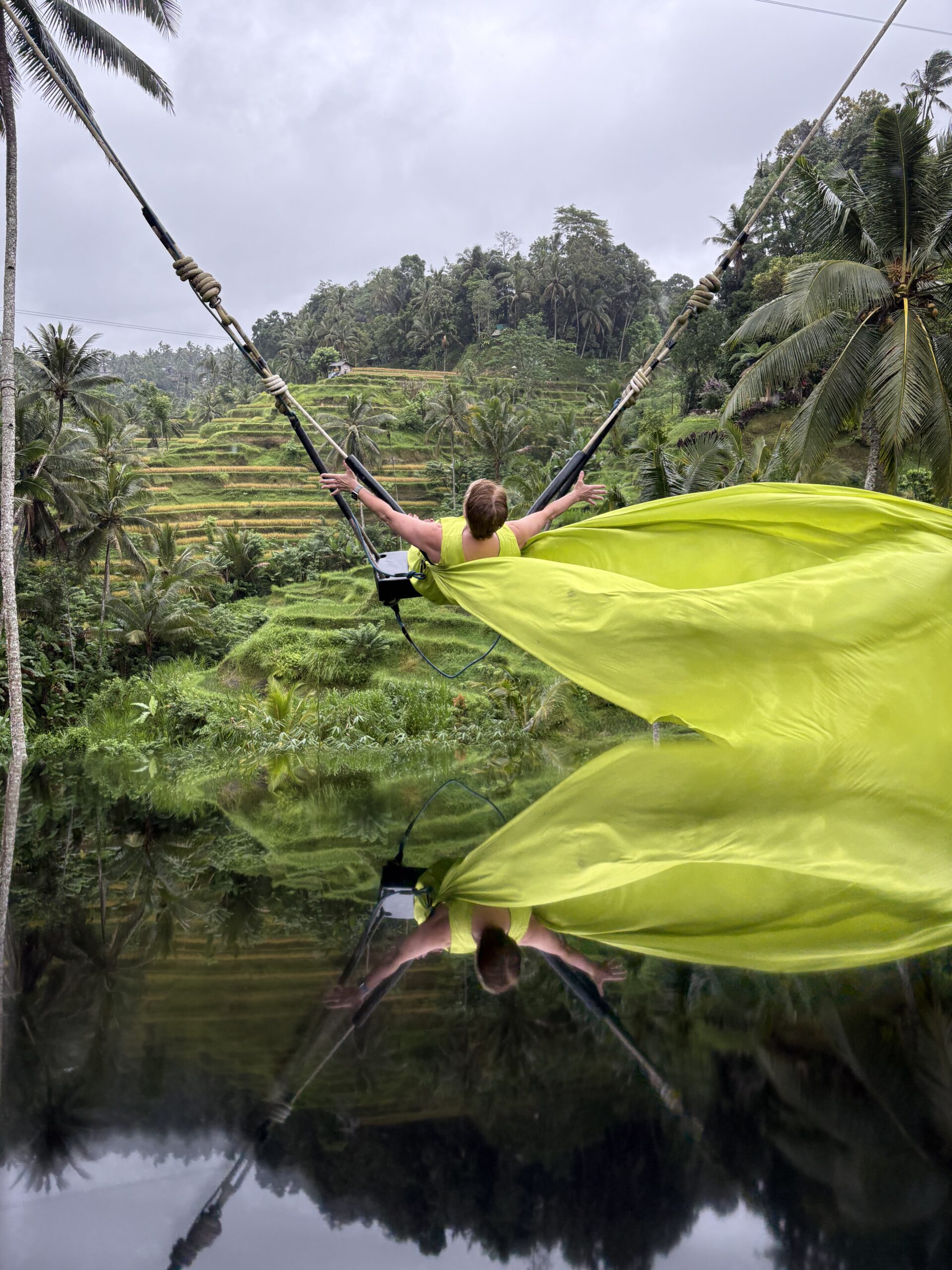 Woman in a flowing yellow-green dress on a swing above a reflection pool, Bali rice terraces behind her