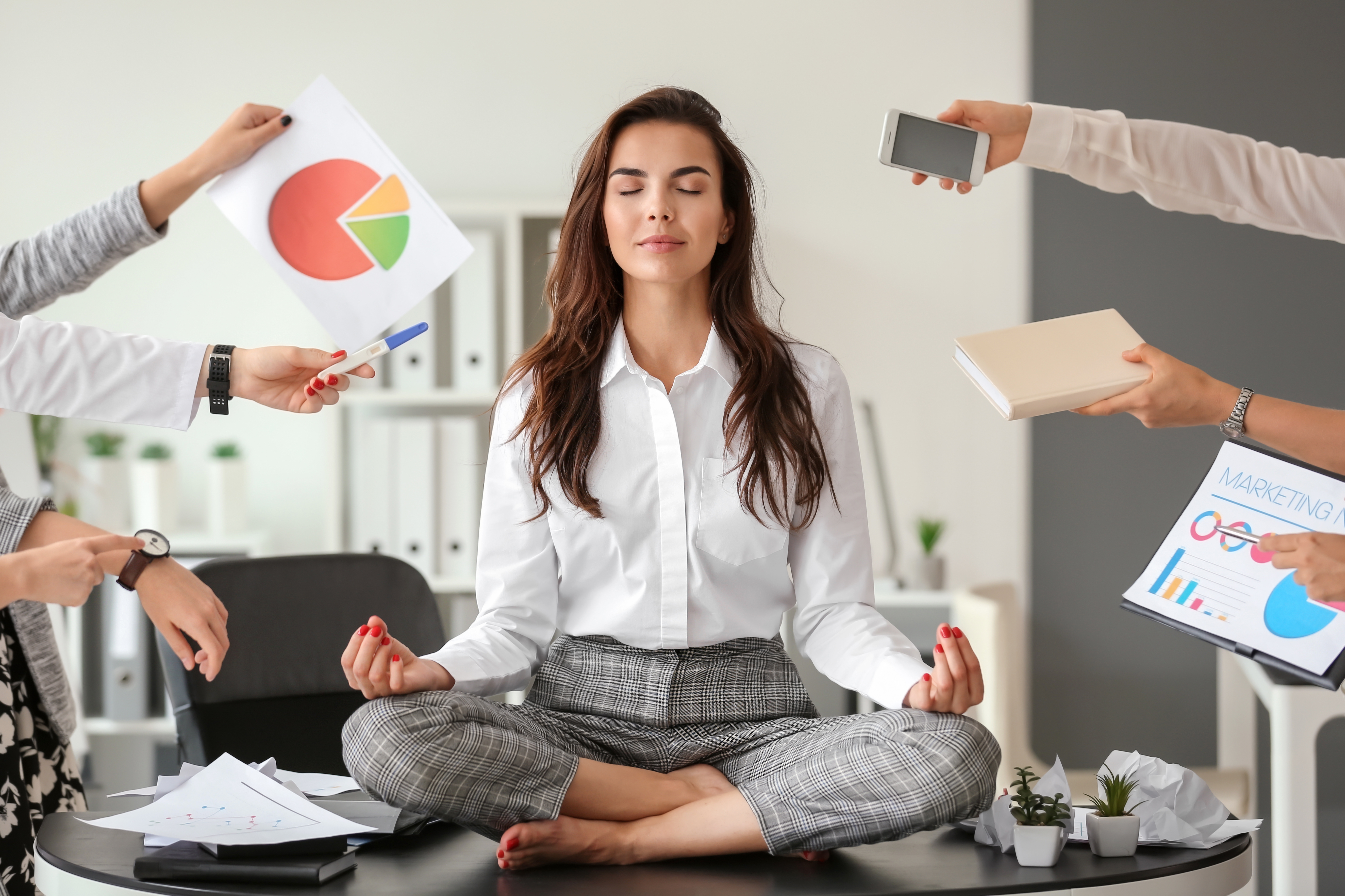 A professional woman sitting calmly in lotus pose on her desk while colleagues push documents, charts and a phone toward her.