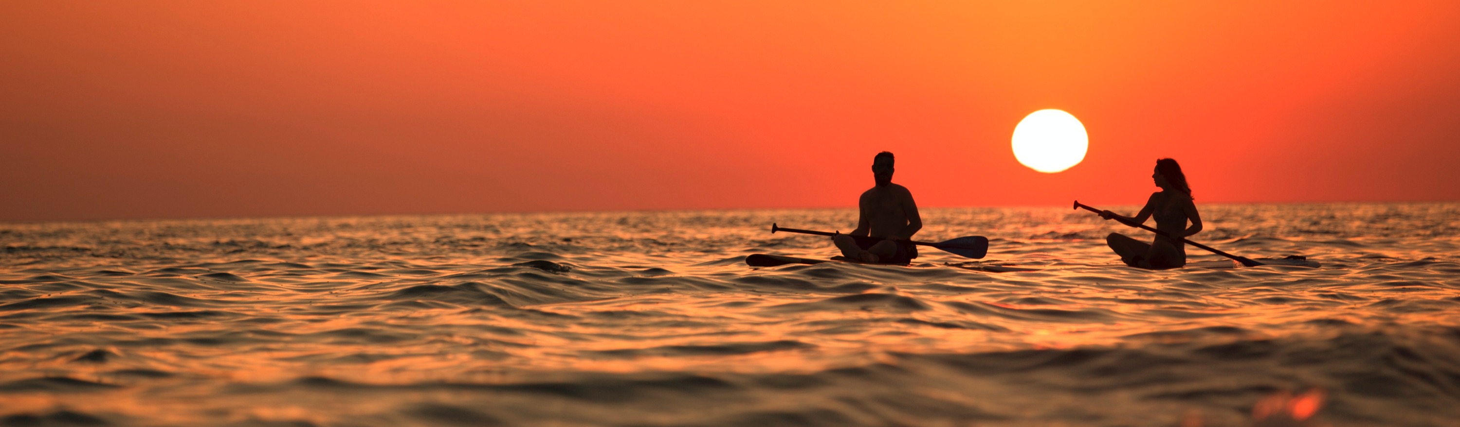 Two people sitting on paddleboards on calm ocean water at sunset.