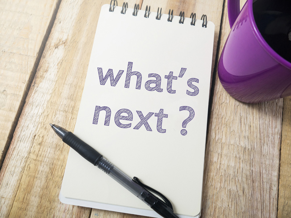 Handwritten note reading 'what's next?' in purple on a wooden desk beside a pen and mug