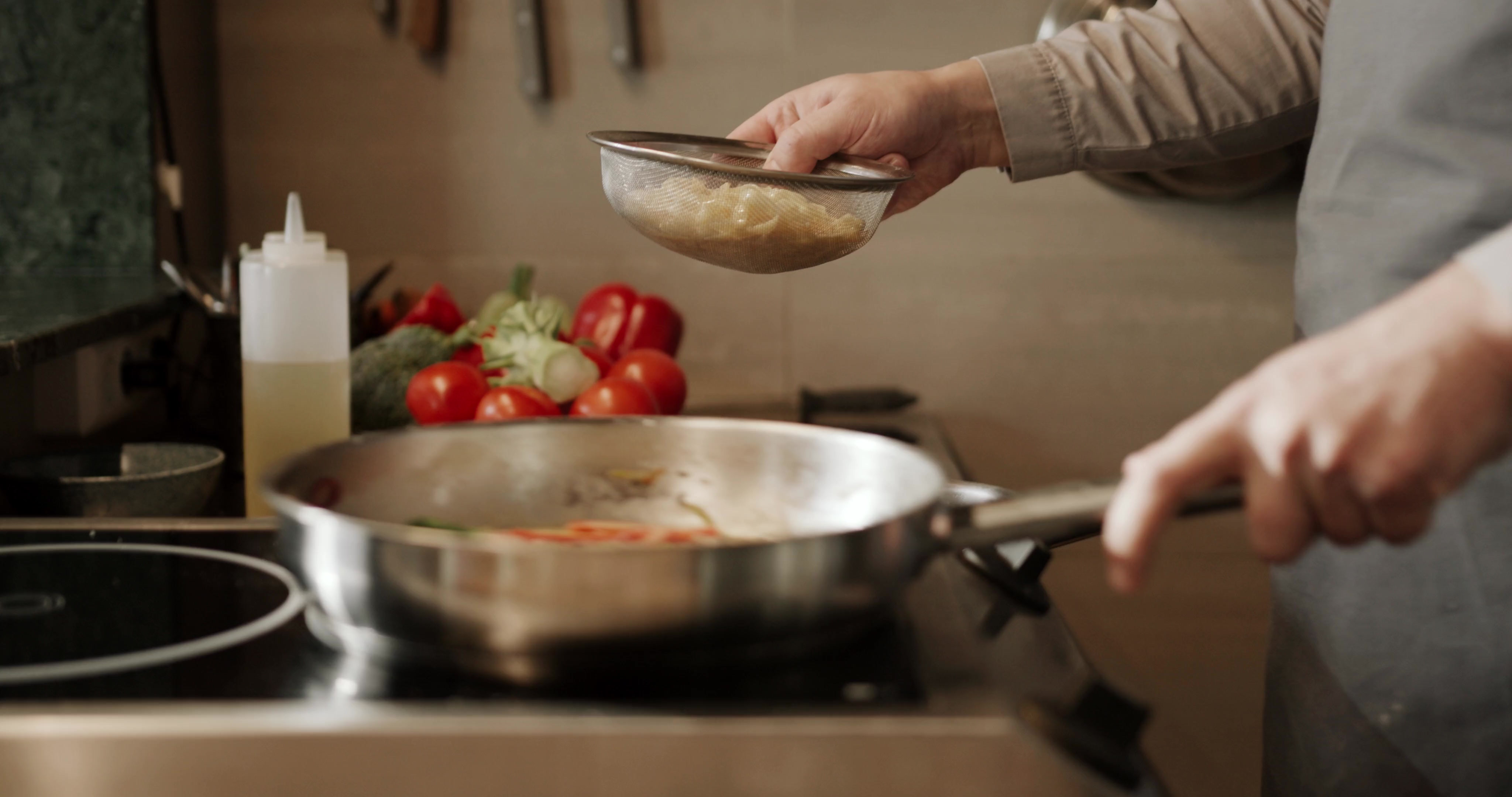 Chef's hands straining pasta into a sauté pan of tomatoes and vegetables