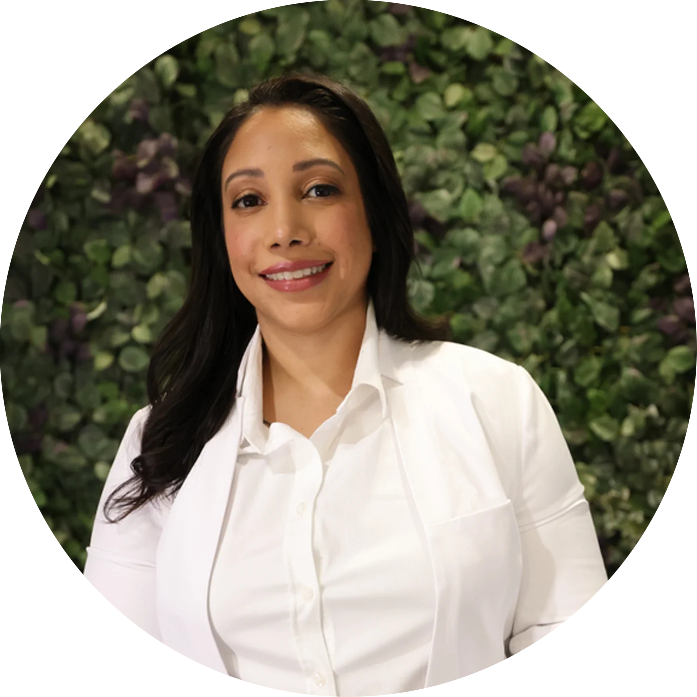 Dr. Neha Pathak, board-certified podiatrist, smiling in front of a green foliage wall.