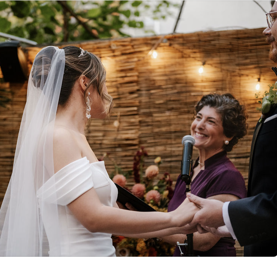 A smiling officiant at a microphone with a bride and groom holding hands at a backyard ceremony with string lights