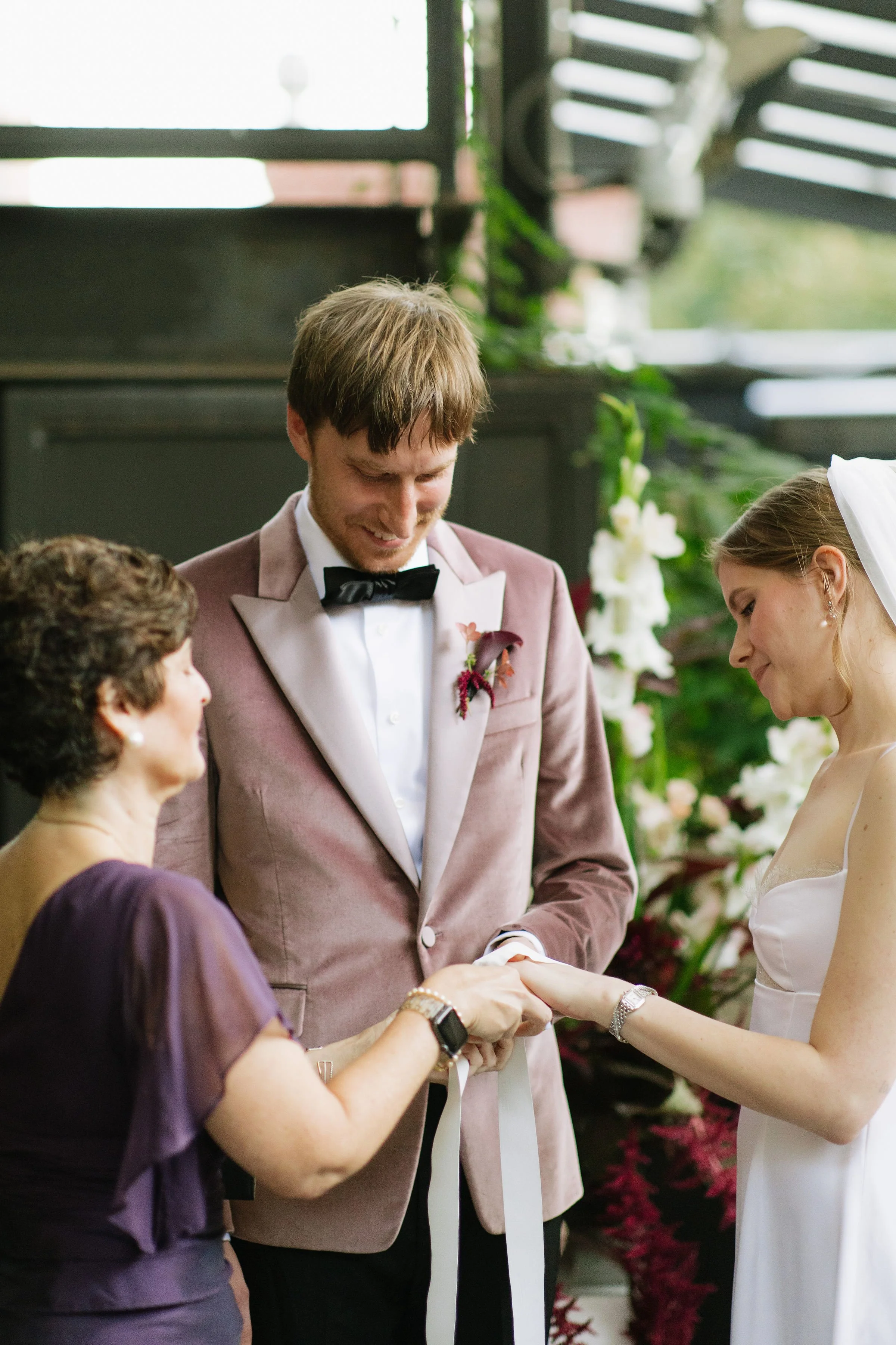 Officiant performing a hand-fasting ritual with a groom in a pink velvet tuxedo and bride in white