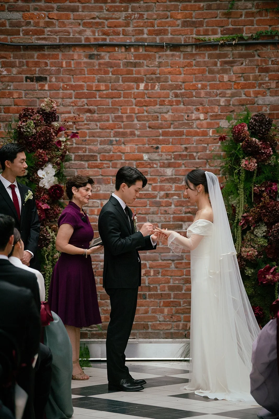 Ring exchange at a brick-wall venue with dark floral arch
