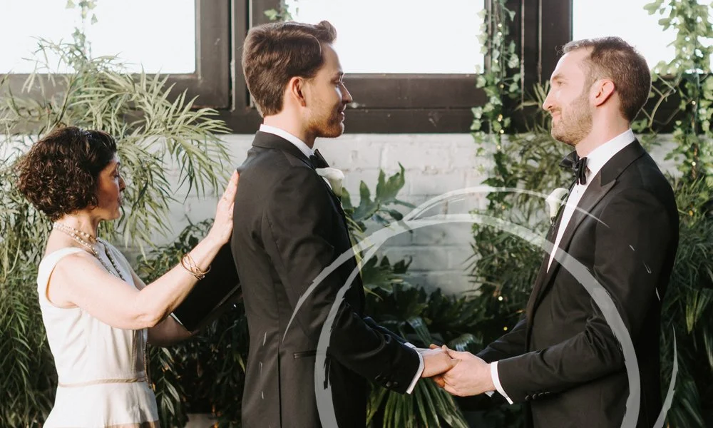 Officiant in white officiating a same-sex male couple holding hands amid tropical plants