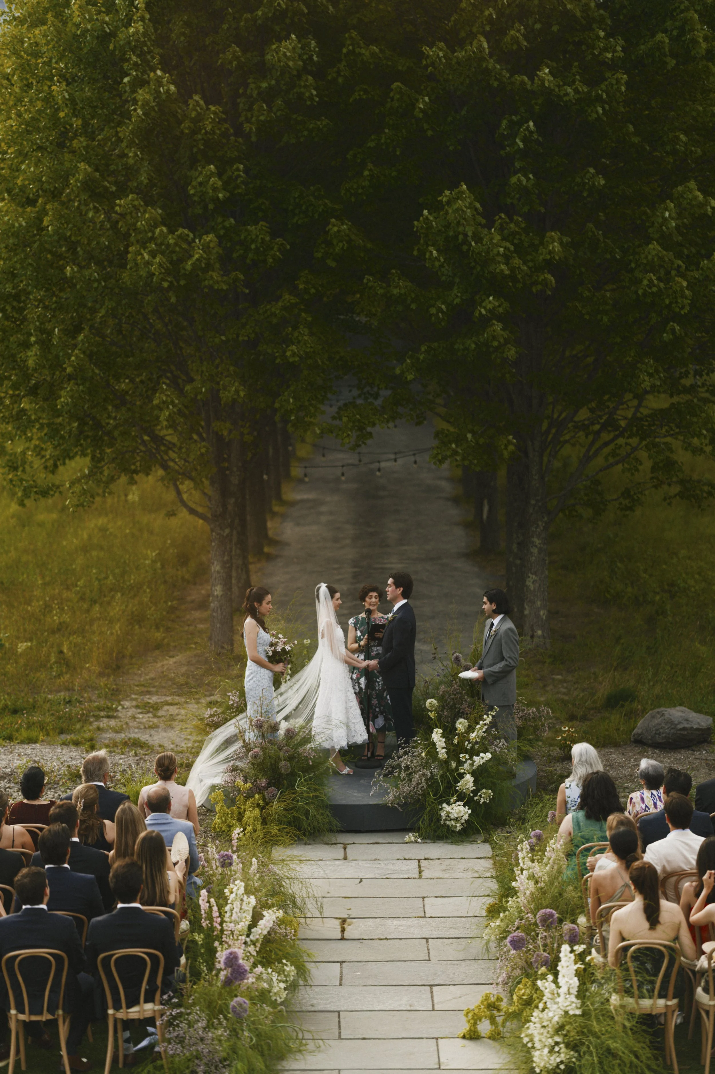 Outdoor wedding ceremony with a couple at the altar, flanked by trees and wildflowers, guests seated along a stone aisle