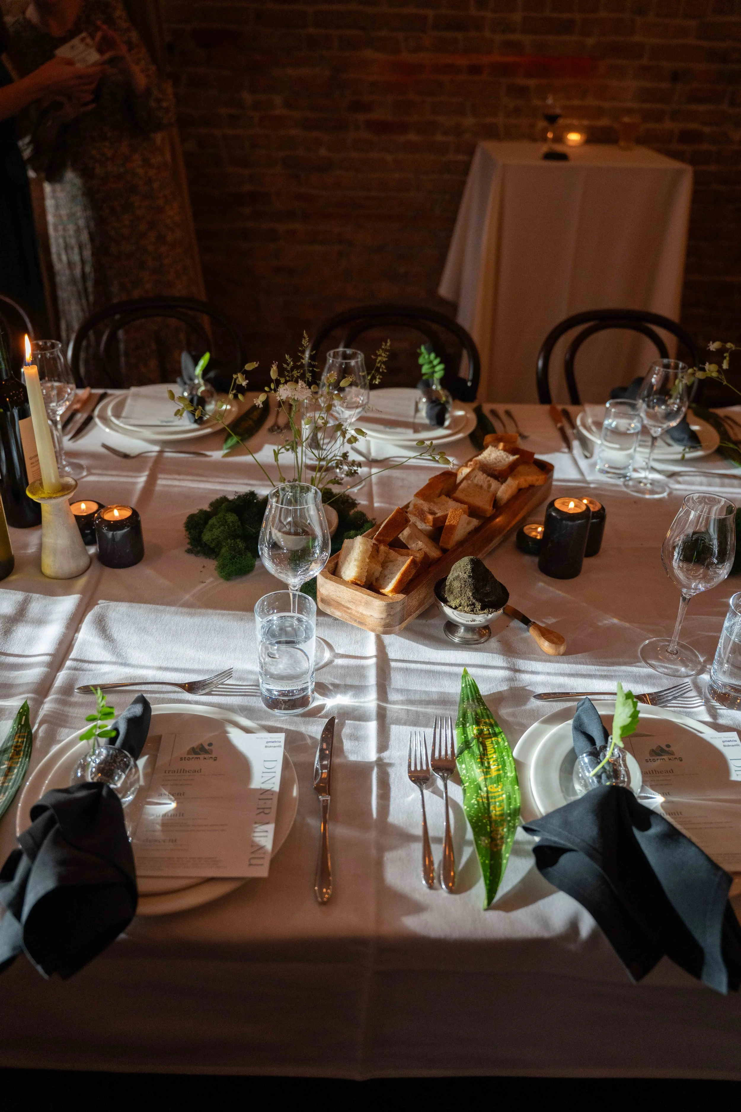 Elegantly set dinner table with black napkins, bread, candles and greenery in a brick-walled venue