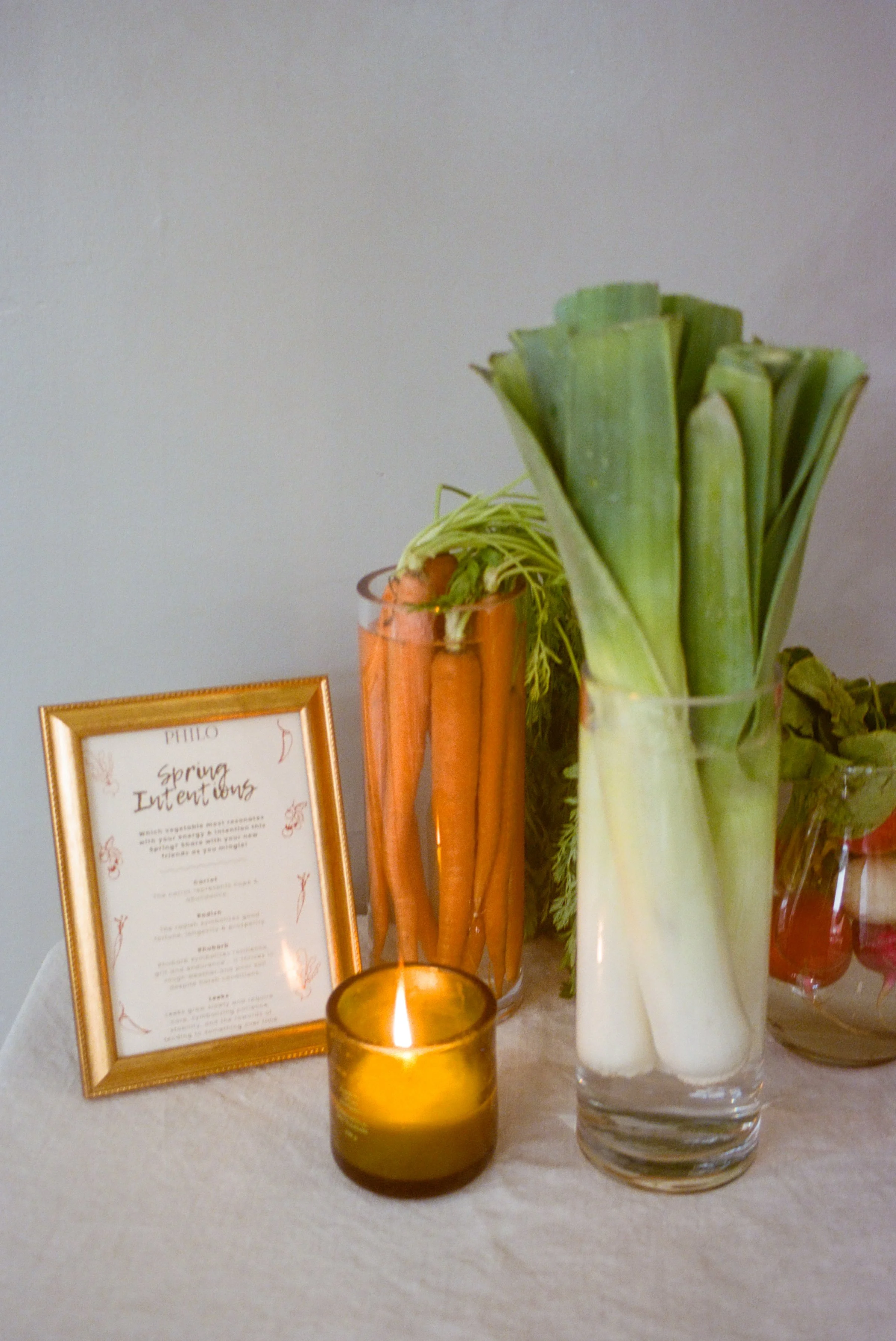 Philo Spring Intentions event menu framed in gold beside leeks, carrots and a lit candle