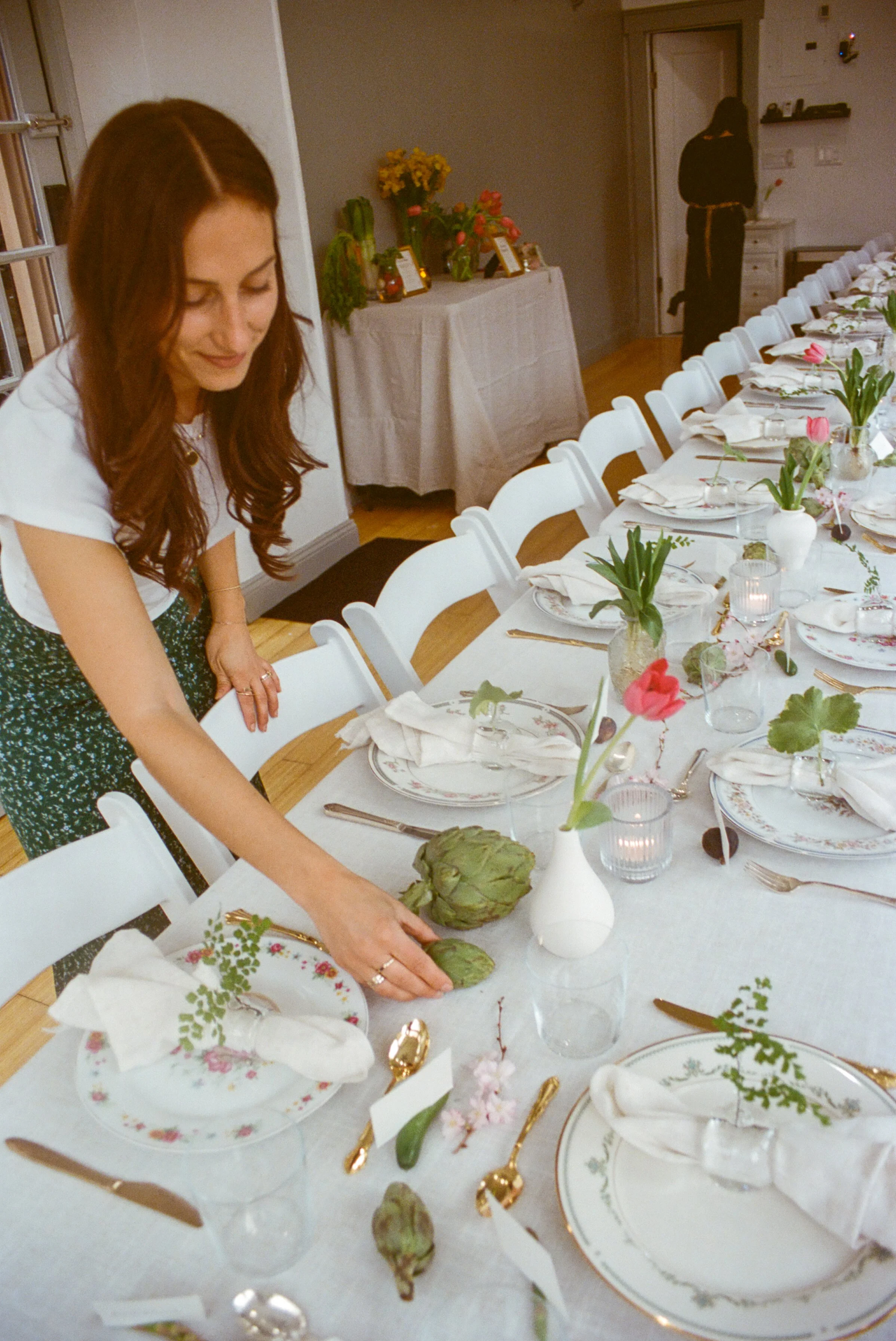 Erica arranging a long spring dinner table with floral china, tulips, artichokes and gold cutlery
