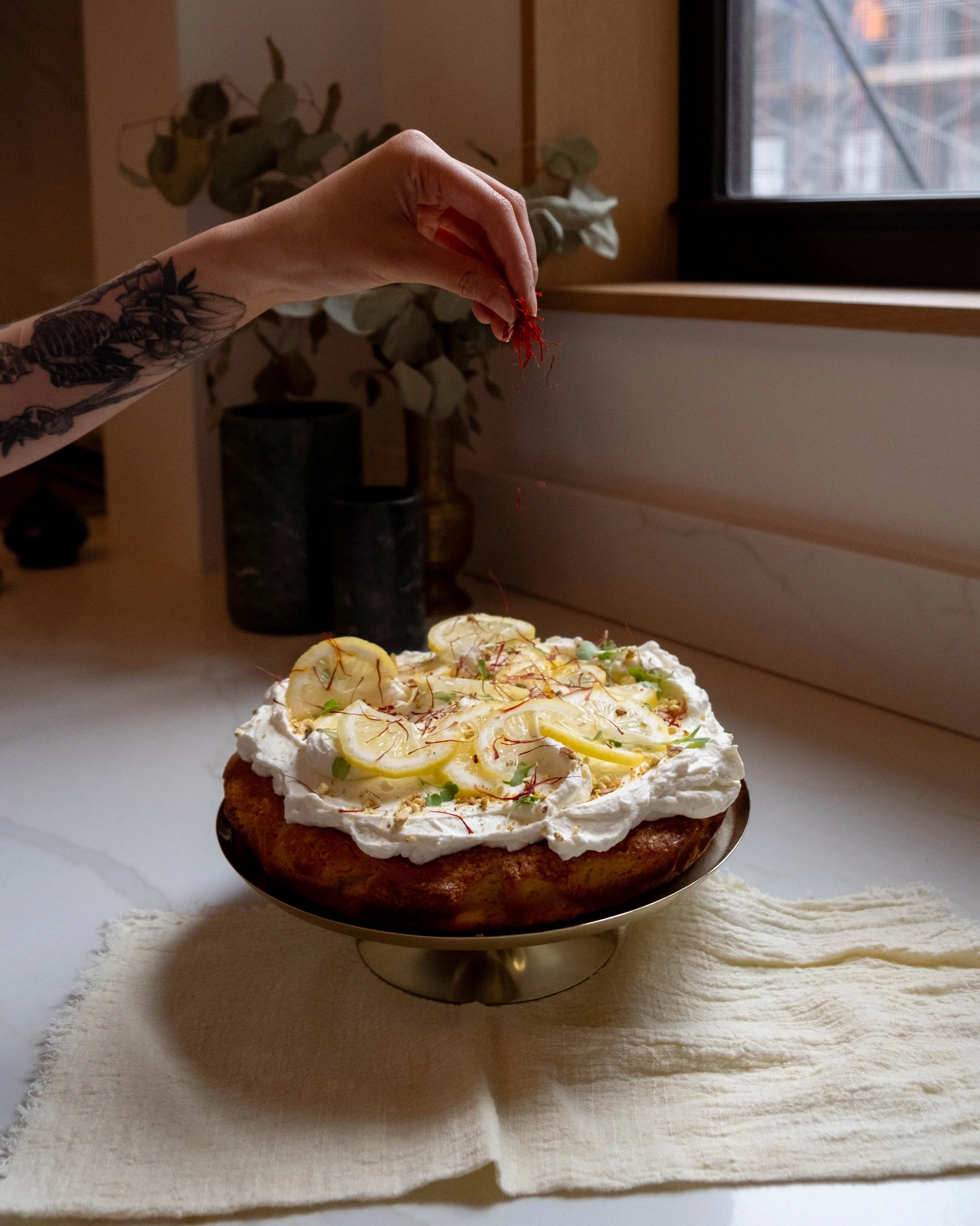 A tattooed hand sprinkling saffron over a lemon cake on a brass cake stand by a window