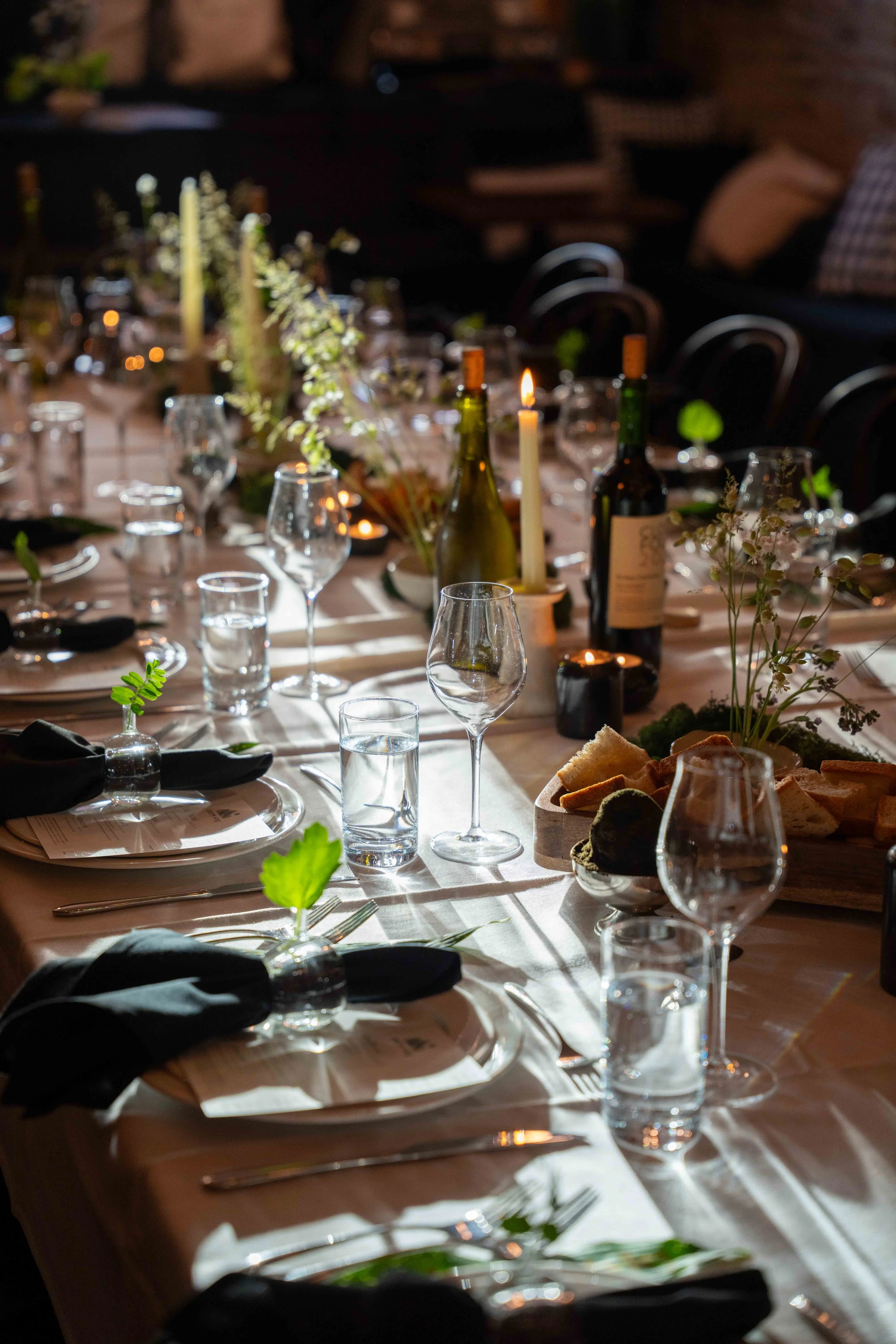 Long candlelit dinner table with wine bottles, black napkins and botanicals in a moody restaurant setting