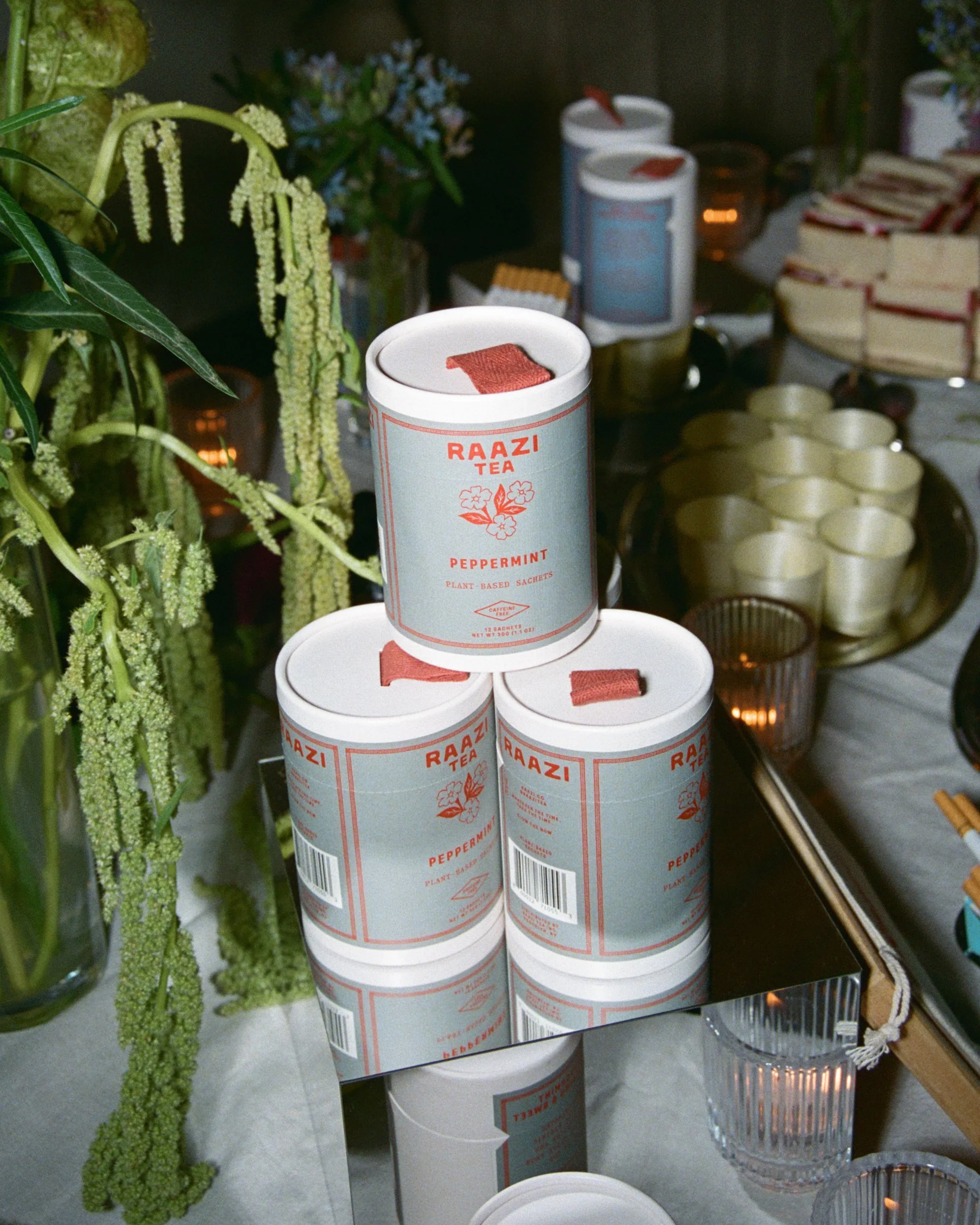Stacked Raazi Tea Peppermint canisters on a mirrored tray with tall green botanicals