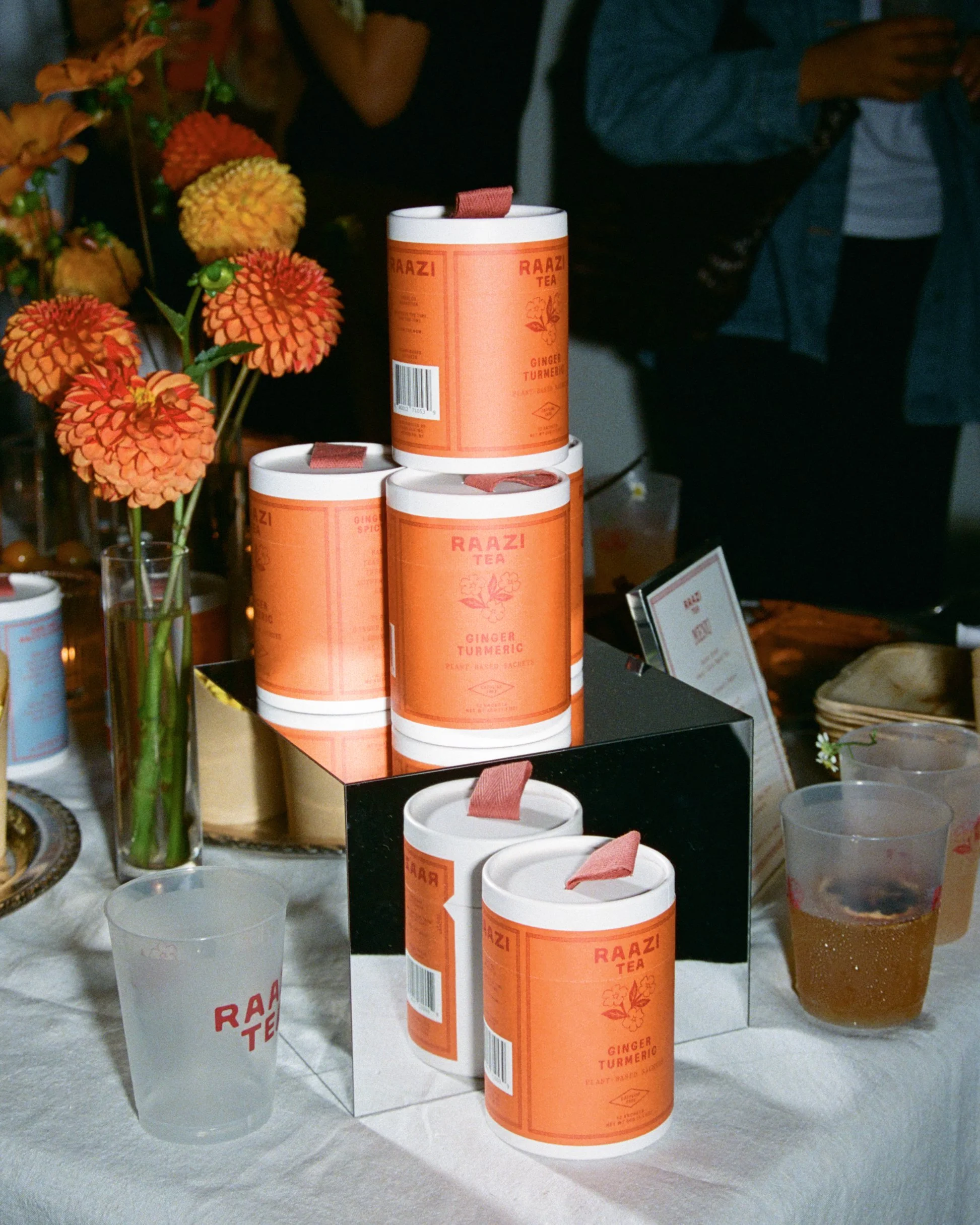 Pyramid of Raazi Tea Ginger Turmeric canisters on a mirrored stand with dahlias