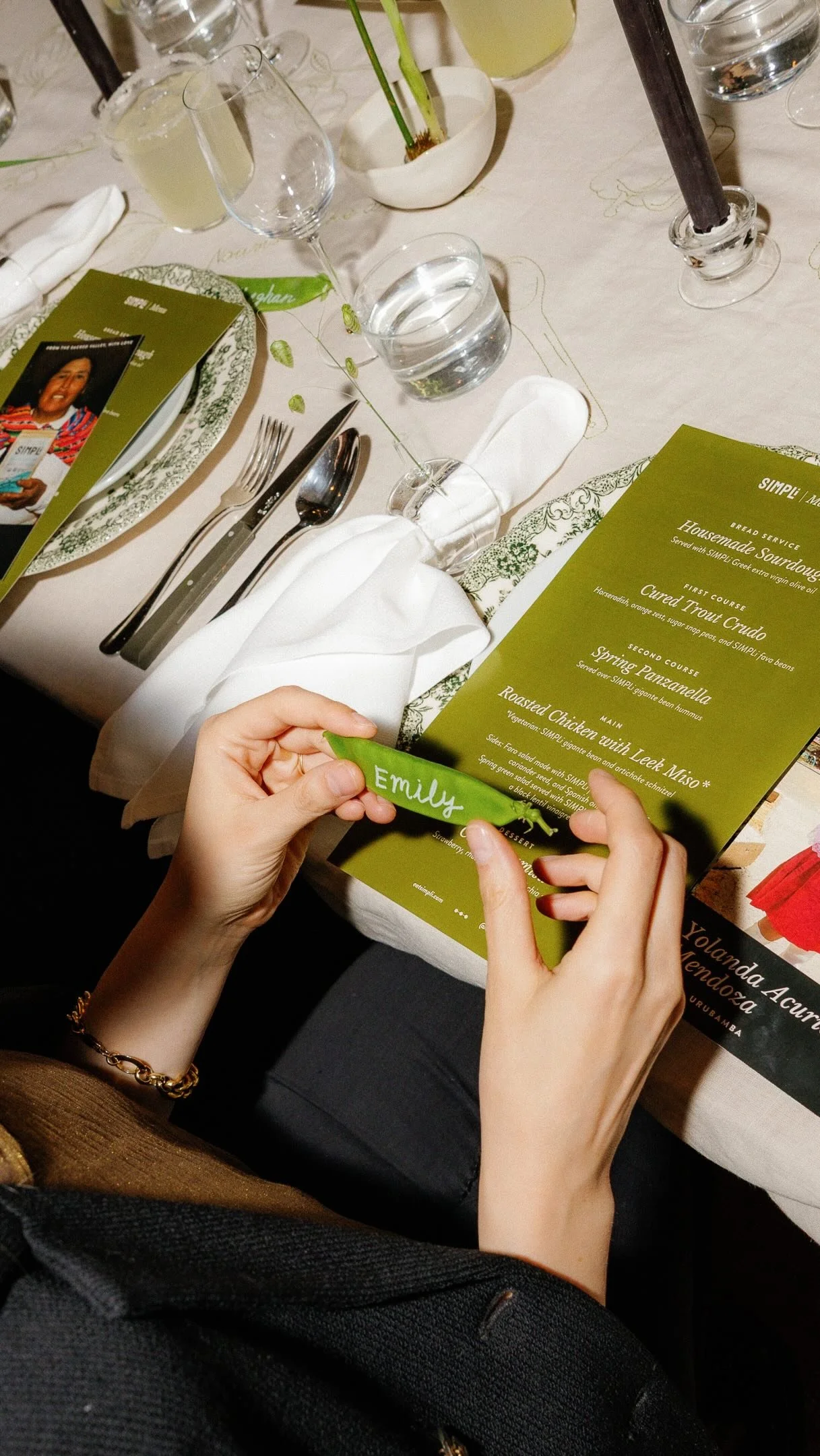 Hands holding a green snap-pea name card over a SIMPLi dinner place setting with an olive-green menu