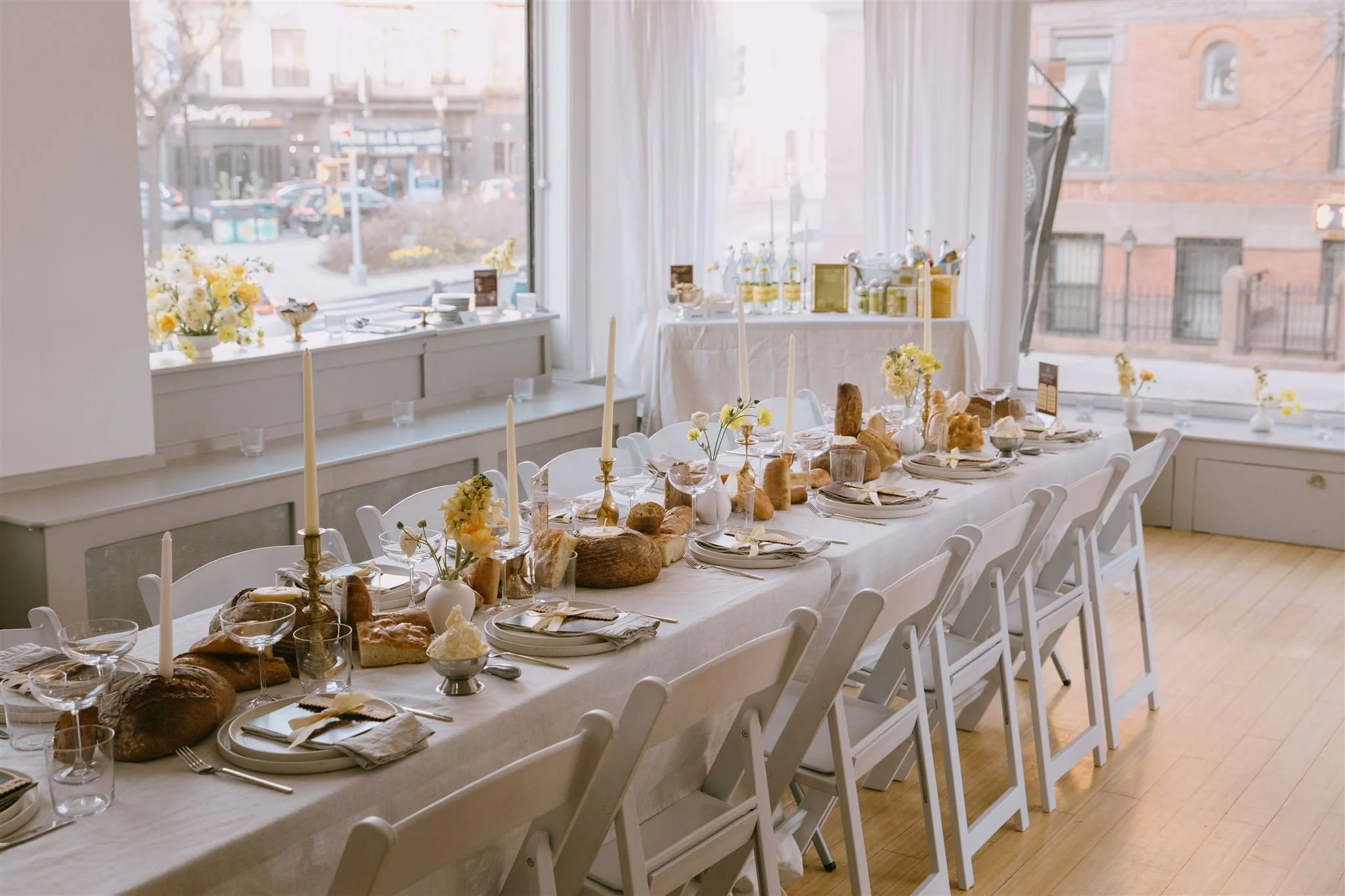 Bright event room with a long white table dressed with bread, taper candles and yellow flowers by tall windows