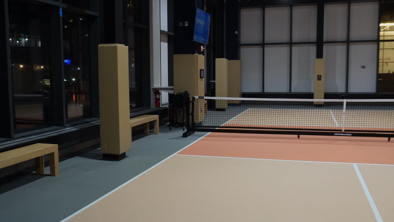 Pickle1's indoor pickleball court at night, with net, coral court surface, wooden benches and floor-to-ceiling windows