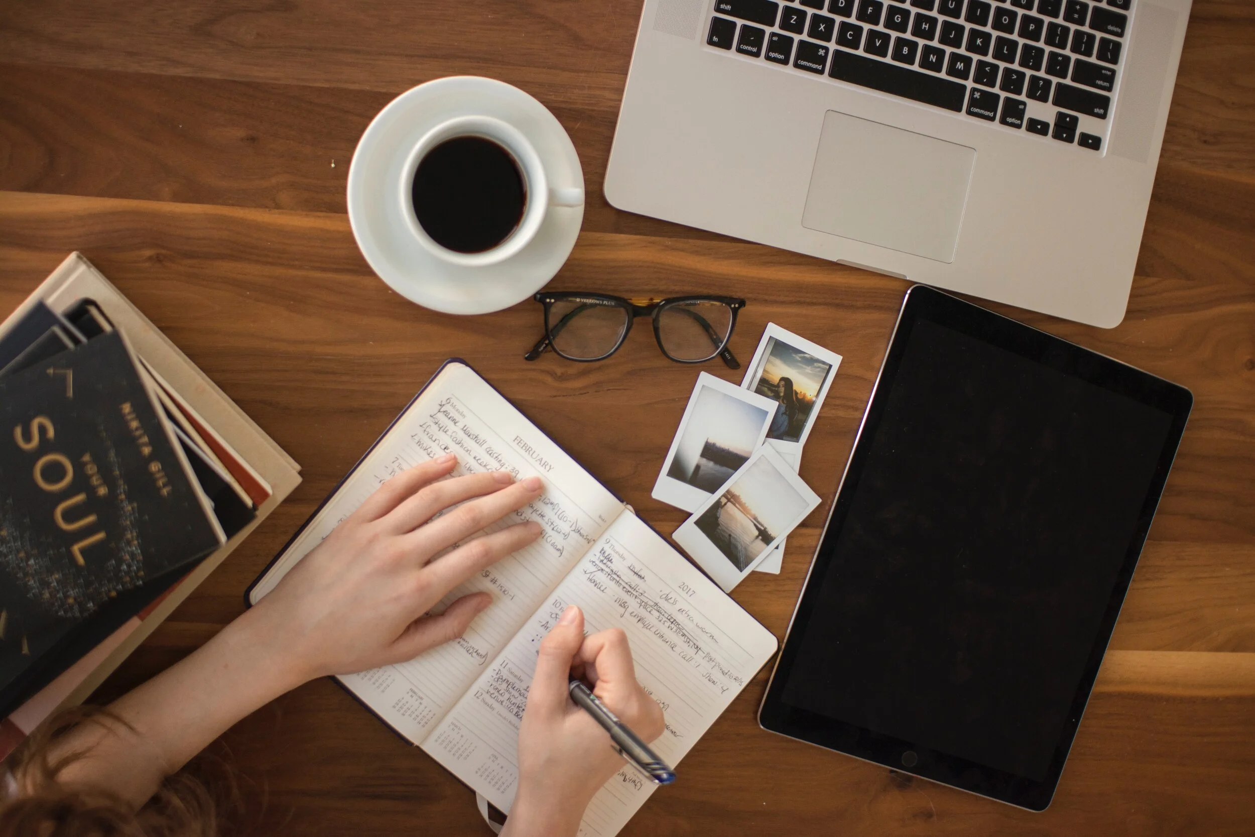 Overhead flat-lay of a wooden desk with a planner, coffee, laptop, tablet, glasses, and polaroid photos
