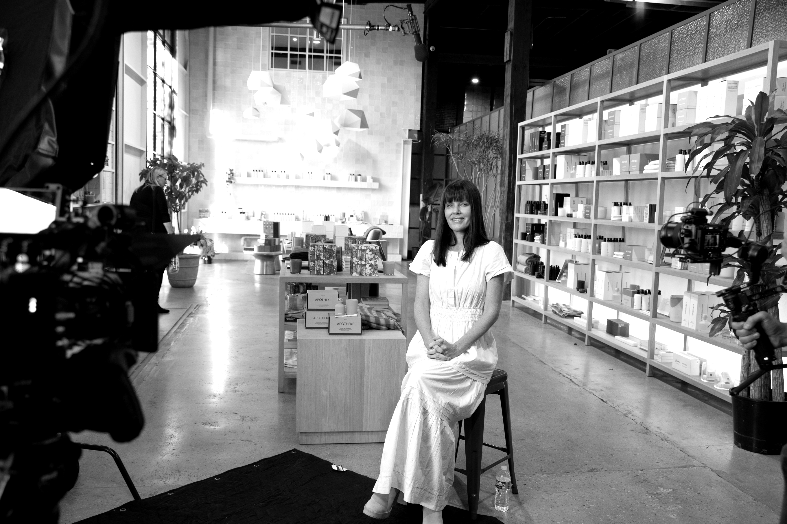 Black-and-white behind-the-scenes shot: a woman in a white dress filmed in an apothecary shop