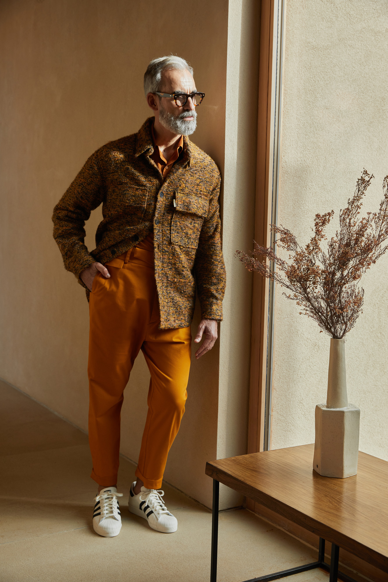 Silver-haired man in a mustard jacket and trousers with Adidas sneakers by a window in a warm interior