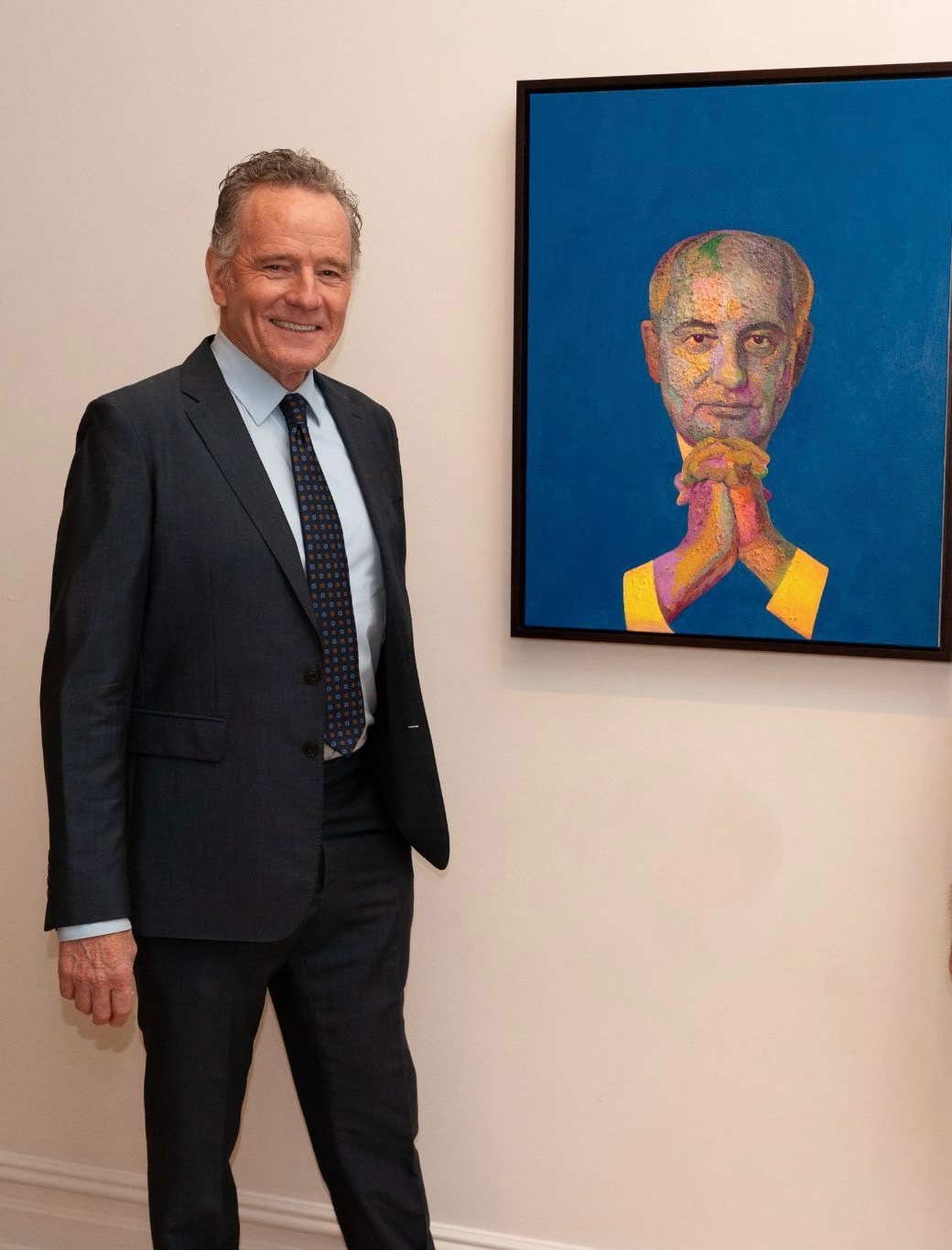 Older man in a dark suit smiling beside a large colourful painted portrait on a gallery wall