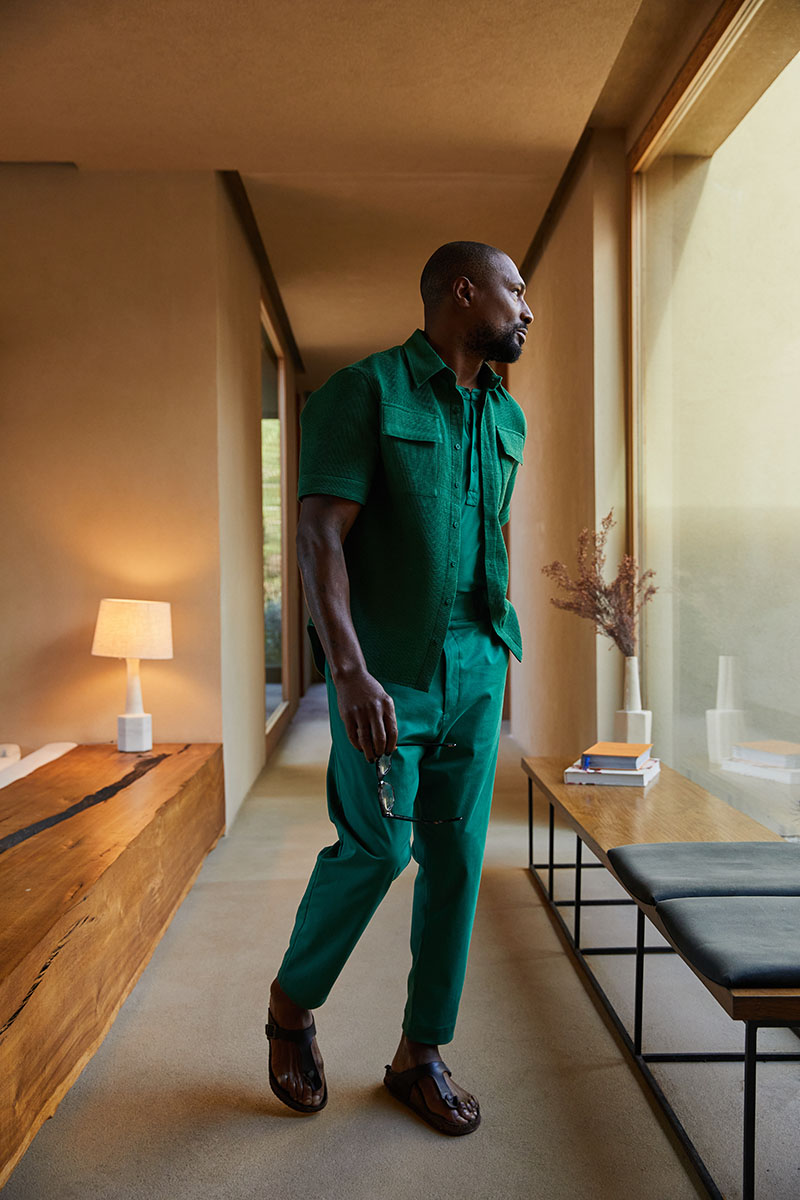Tall Black man in a full emerald-green outfit walking through a warm modern hallway
