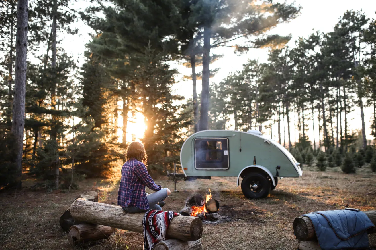 Person sitting by a campfire on logs in a pine forest at sunset with a vintage teardrop camper behind