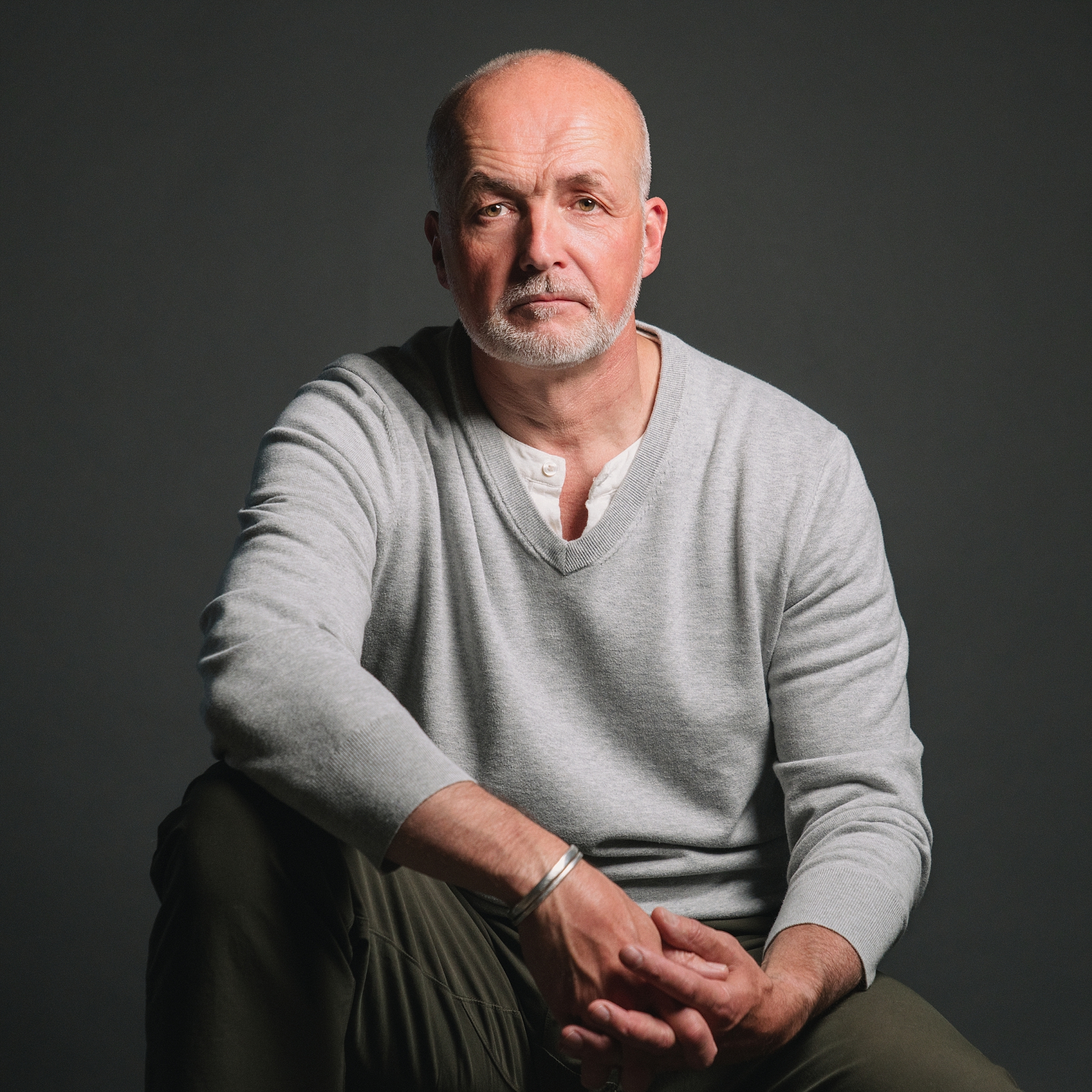 Middle-aged bald man with grey stubble in a grey V-neck sweater, dramatic studio lighting on a dark grey background