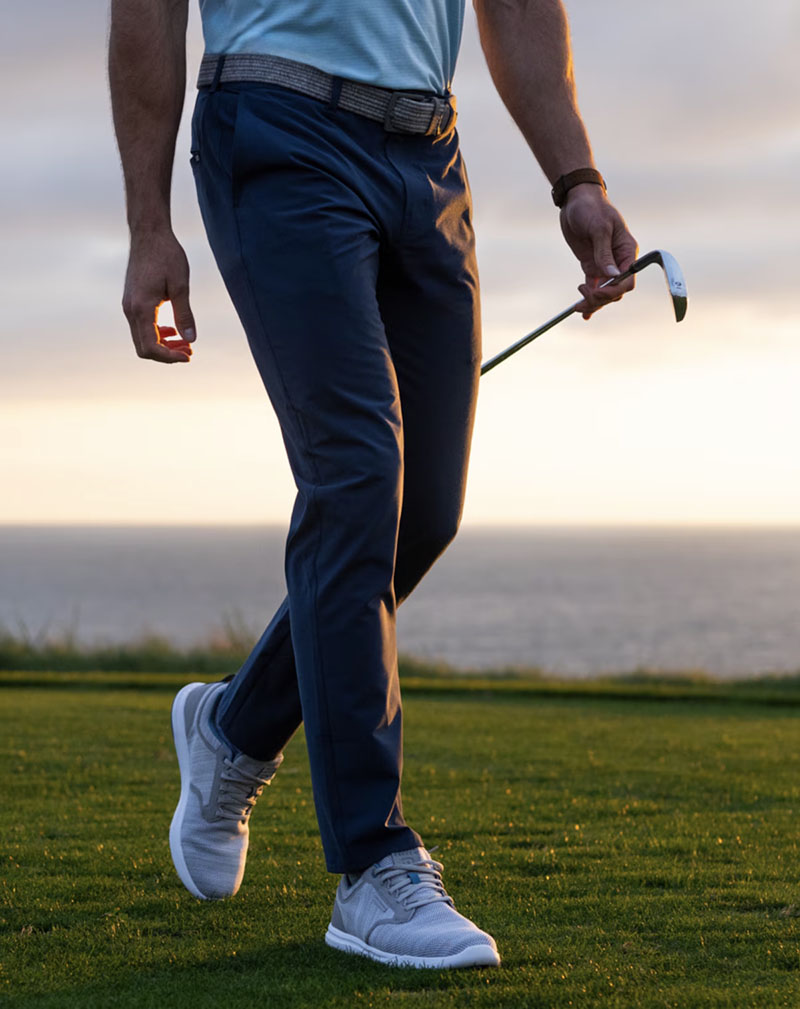 Man in navy golf trousers holding a putter on a course with an ocean backdrop at sunset