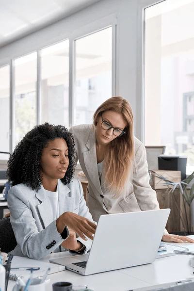 Two businesswomen in a bright office, one seated pointing at a laptop and one standing with glasses