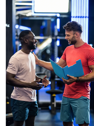 Two men in a gym, one in a red shirt holding a clipboard, consulting with a client
