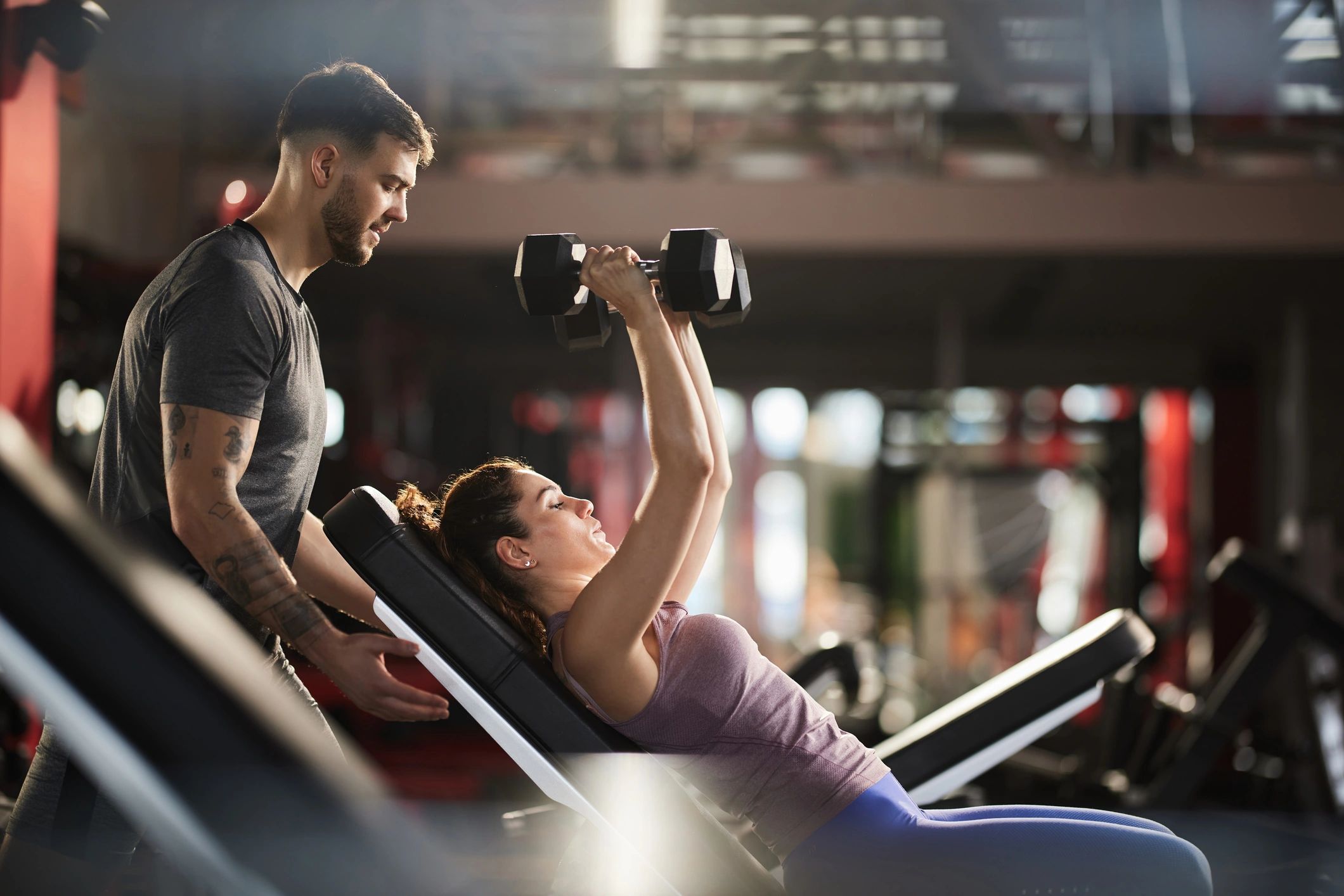 Male personal trainer spotting a woman pressing dumbbells on an incline bench in a gym