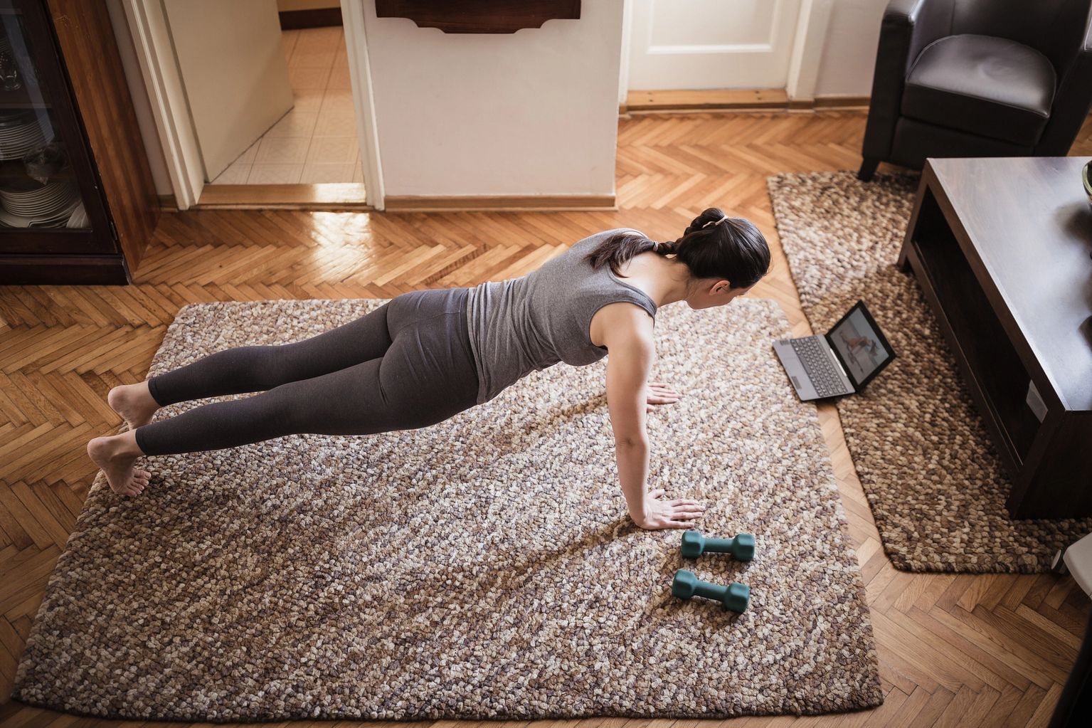 Woman holding a plank on a rug at home with dumbbells nearby and a laptop open beside her