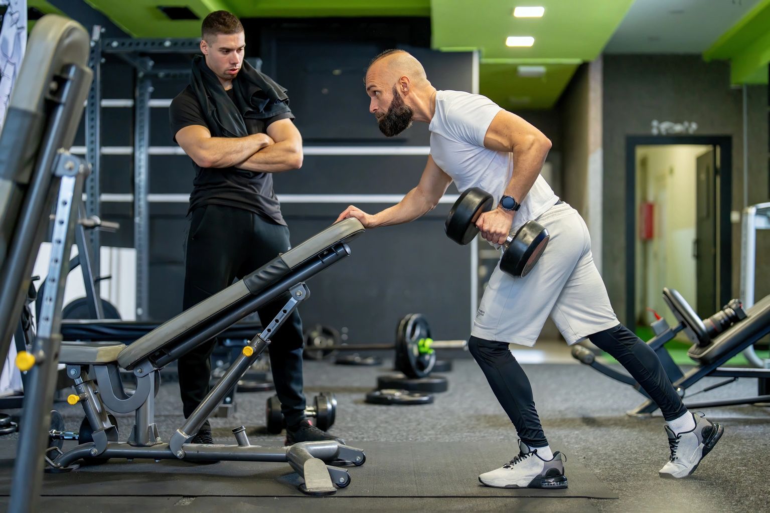 Man performing a single-arm dumbbell row while a trainer watches his form in a green-accented gym