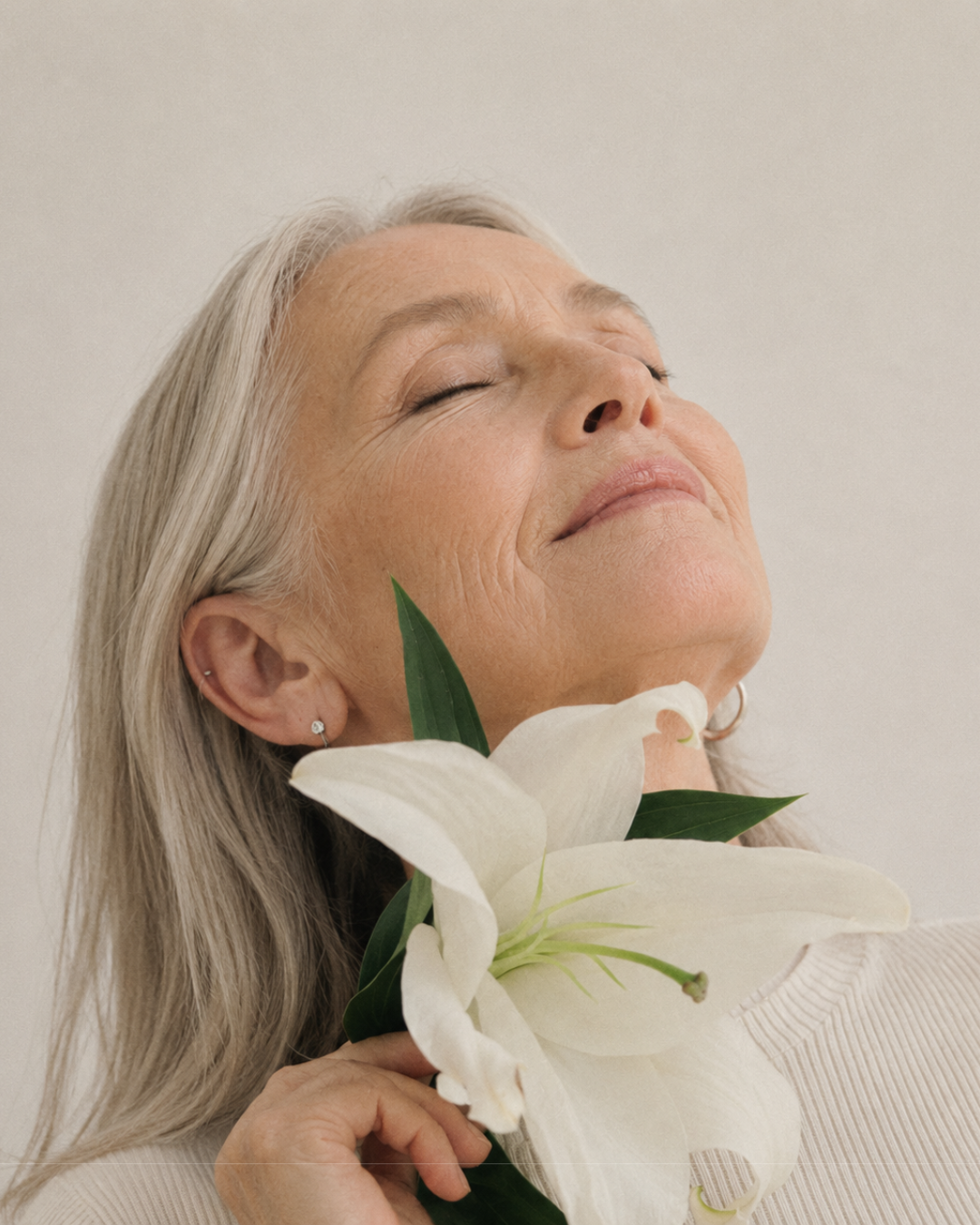 An older woman with long gray hair, eyes closed, holding a white lily flower in a moment of calm