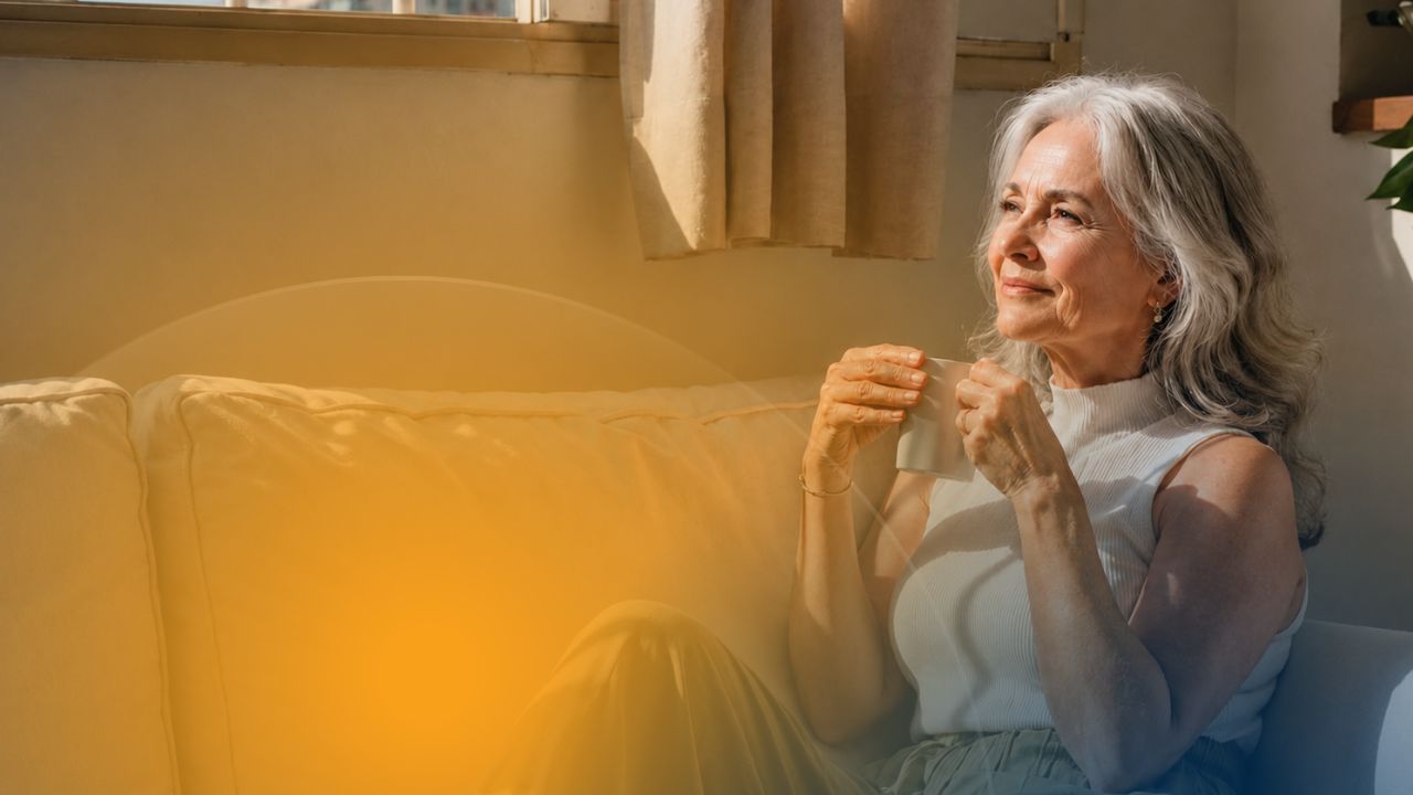 A silver-haired woman holding a cup of tea, gazing thoughtfully out a sunlit window