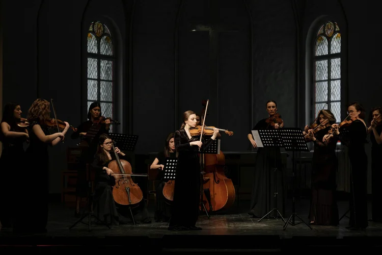 Violinist soloist performing at center stage with a string orchestra in a dark gothic hall.