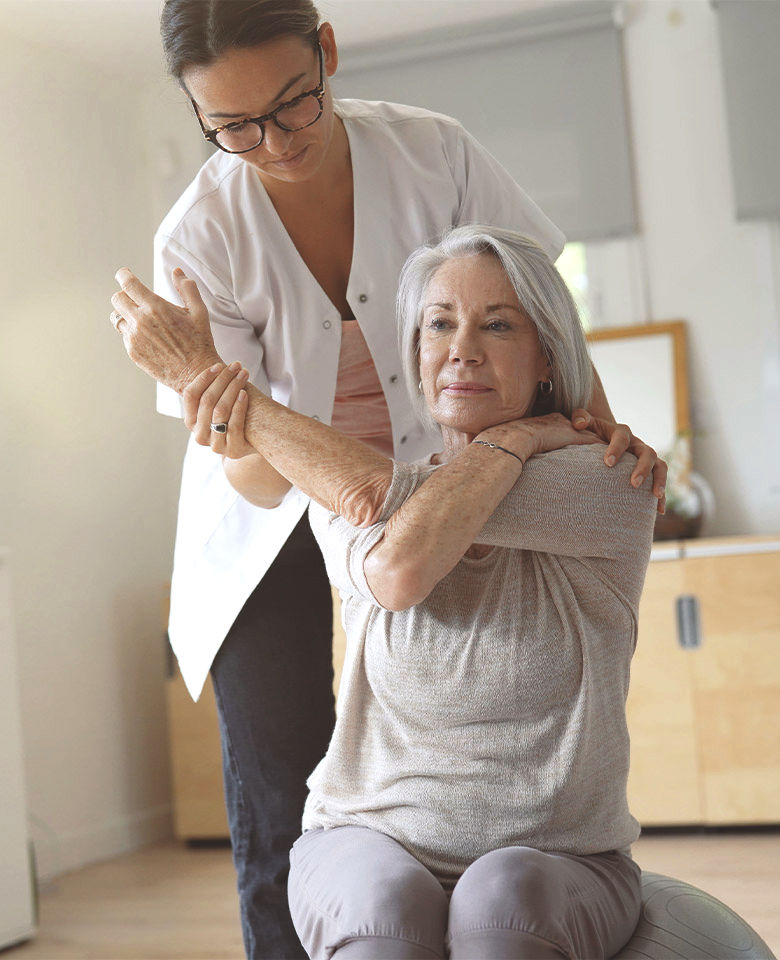 Physical therapist in a white coat assisting an older woman with a shoulder stretch on a stability ball