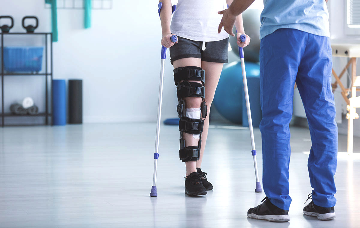 Patient with a knee brace on crutches being assisted by a therapist