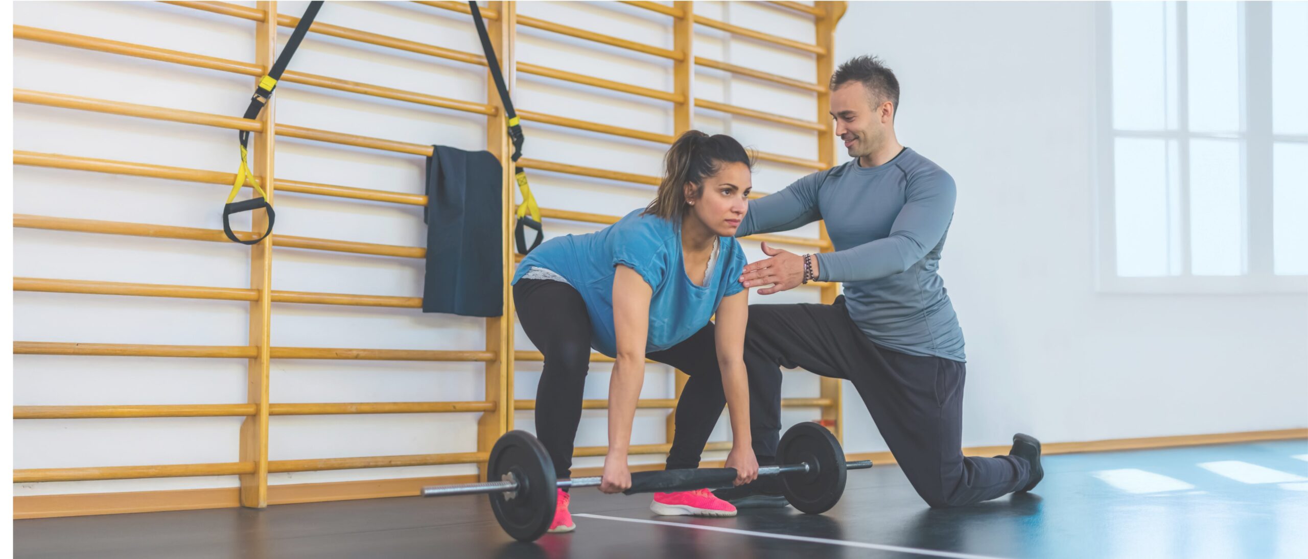 Personal trainer coaching a woman deadlifting a barbell in a gym with wall bars
