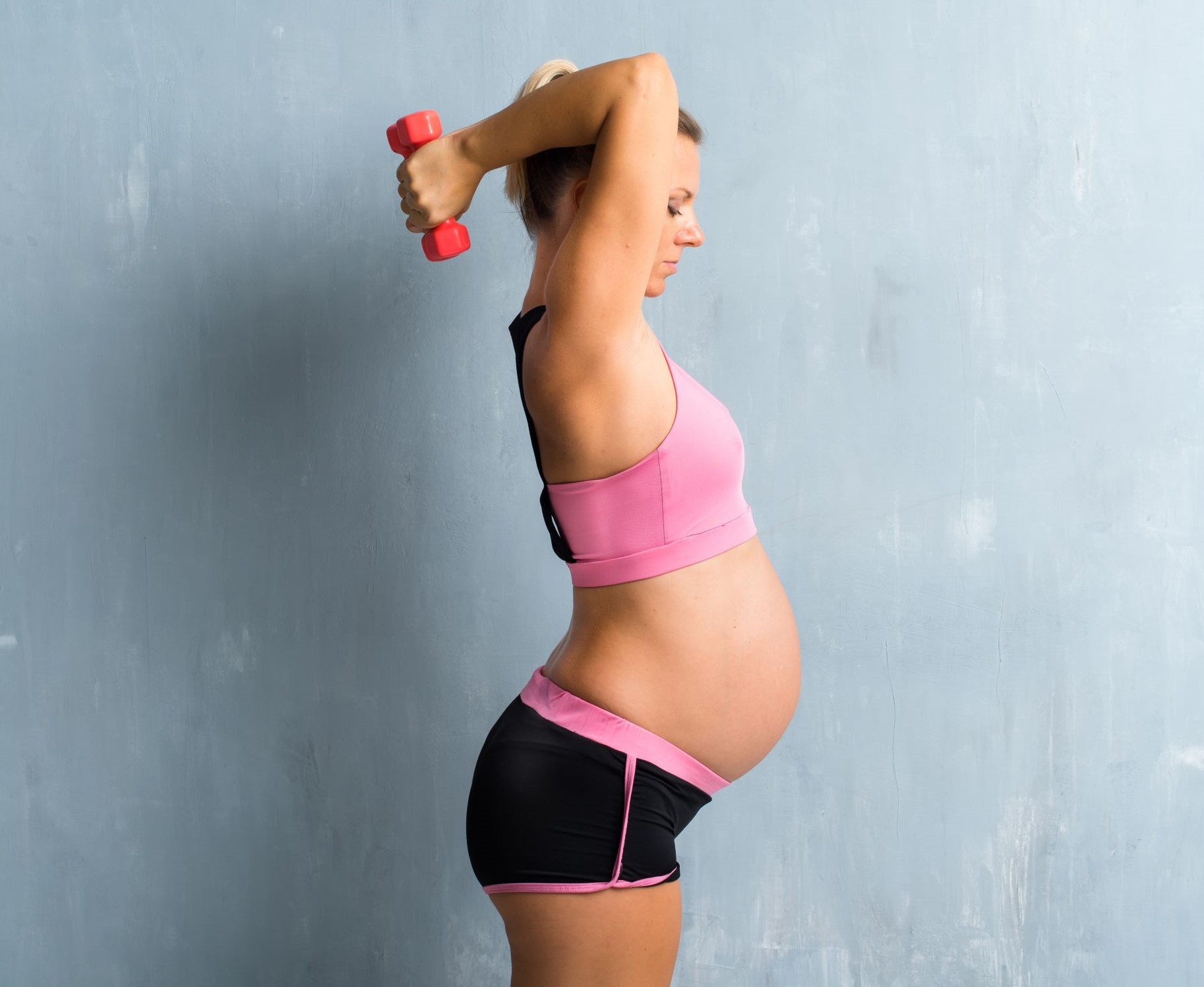 Pregnant woman holding a dumbbell overhead against a grey wall