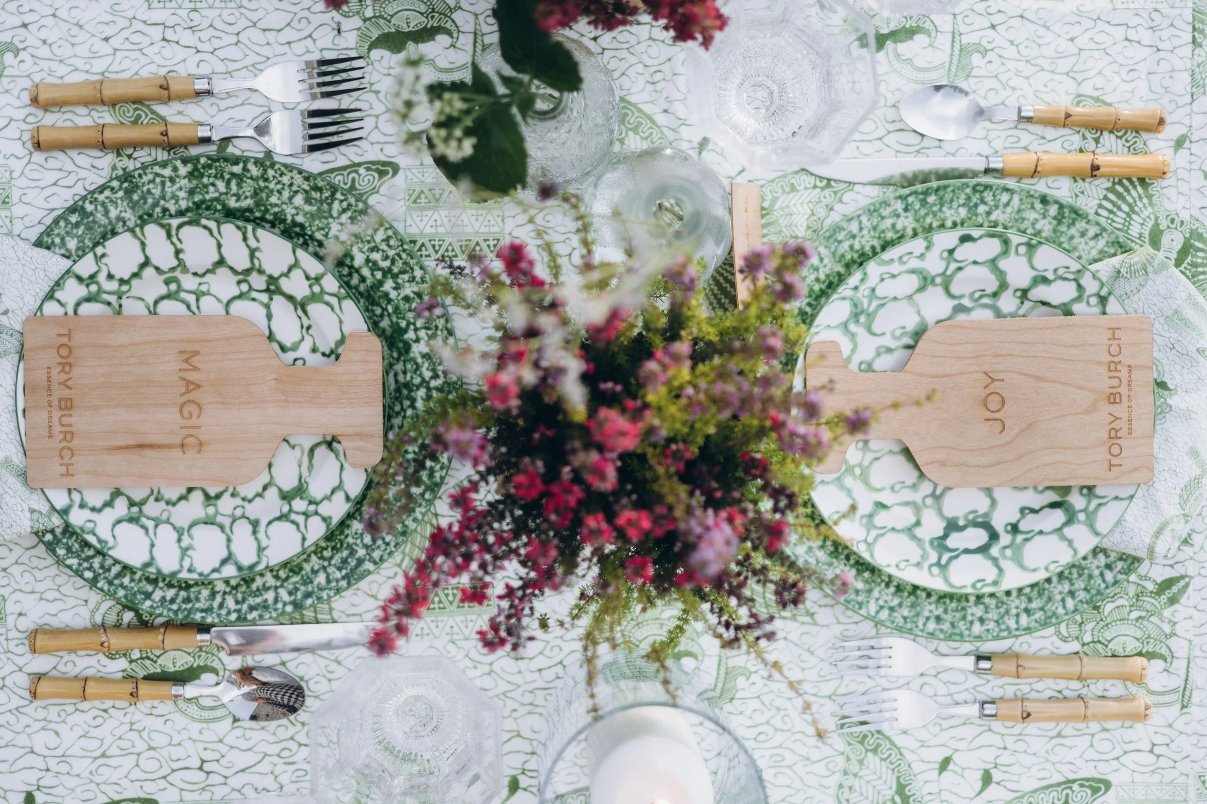 Overhead shot of two Tory Burch Essence of Dreams place settings with wooden menus and green patterned plates