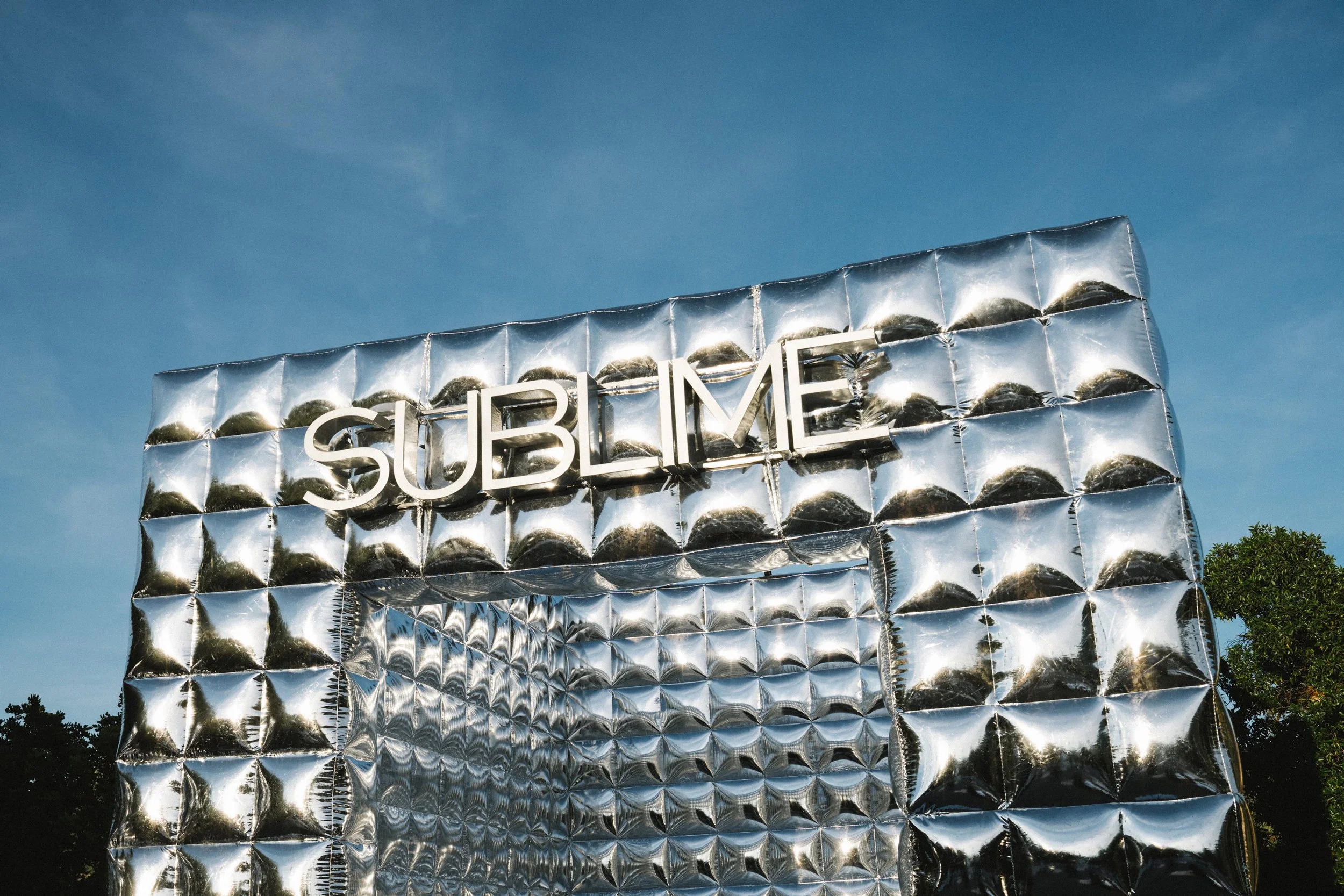 Large silver mylar balloon wall installation reading SUBLIME against a blue sky, built for Tory Burch in Mallorca