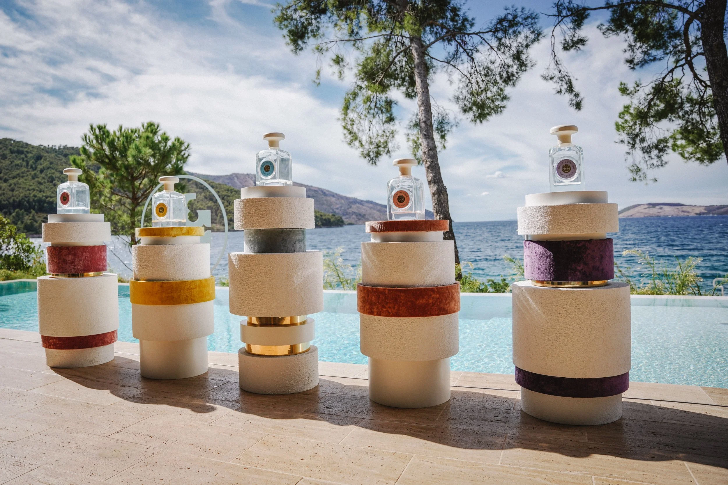 Four Tory Burch Essence of Dreams fragrance bottles on stacked sculptural pedestals beside a pool